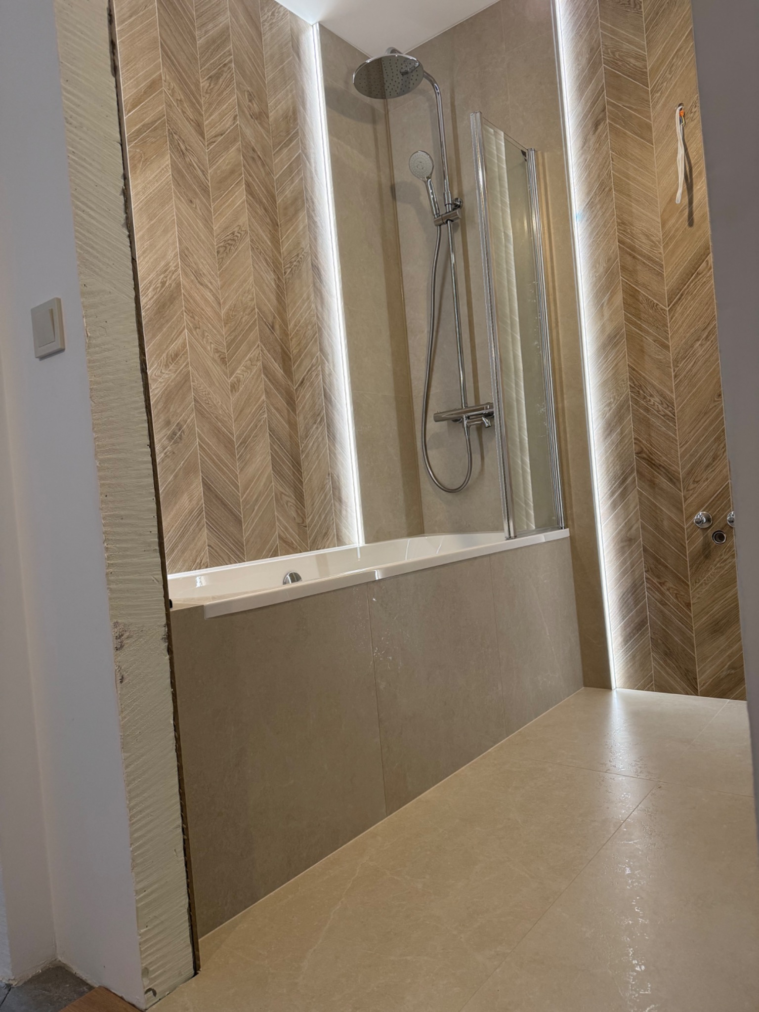 Wykończona łazienka z beżowymi płytkami ułożonymi w jodełkę, podświetleniem LED i kabiną prysznicową. Widoczna wanna i armatura. Całość w ciepłej tonacji.