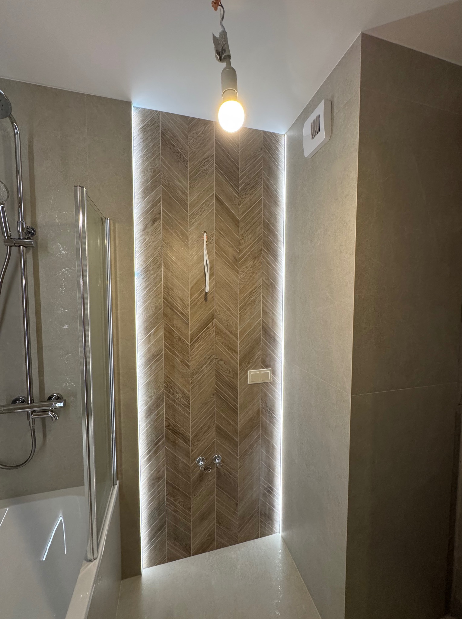 Nowoczesna łazienka z kabiną prysznicową wyłożoną płytkami w jodełkę, podświetlona listwą LED. Widoczna bateria prysznicowa i wentylator.
