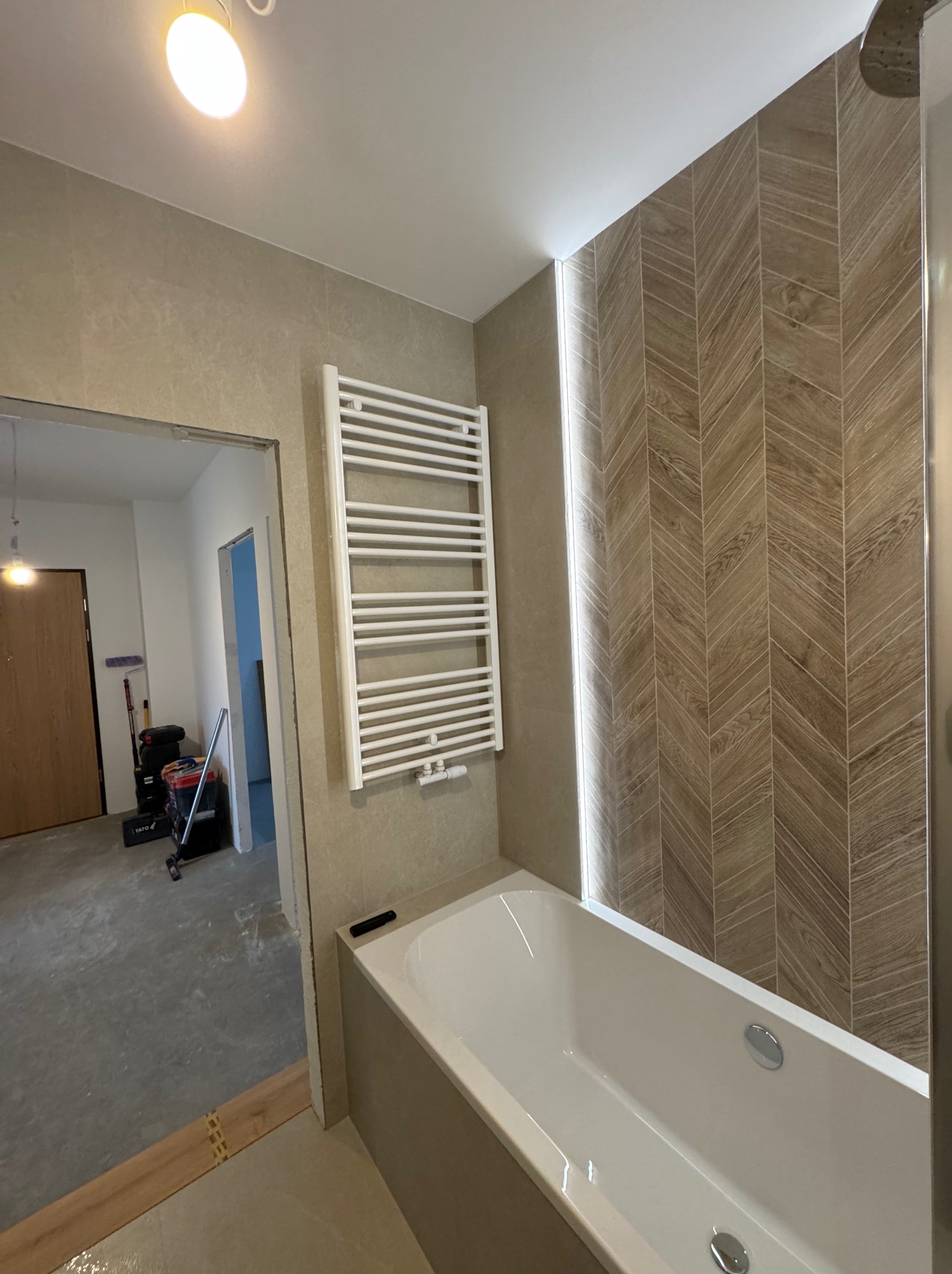 Nowoczesna łazienka z wanną, grzejnikiem drabinkowym i płytkami w jodełkę. Widoczny fragment otwartego przejścia do innego pomieszczenia z narzędziami.