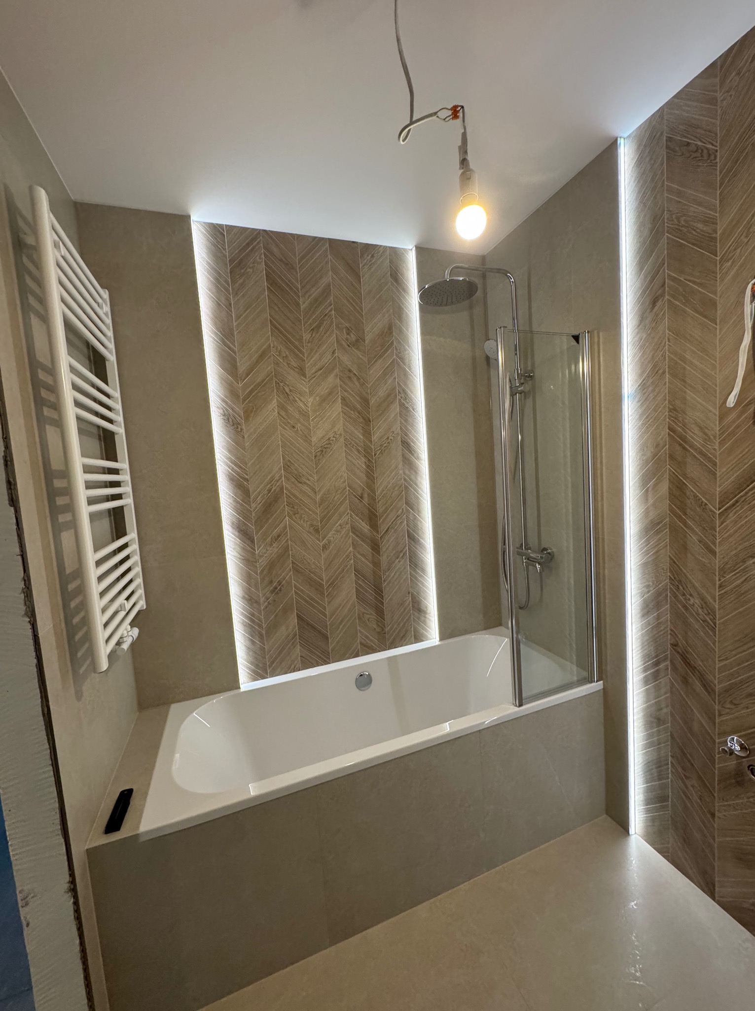 Nowoczesna łazienka z wanną obudowaną płytkami, szklanym parawanem, grzejnikiem drabinkowym i oświetleniem LED wzdłuż ścian z wzorem jodełki.