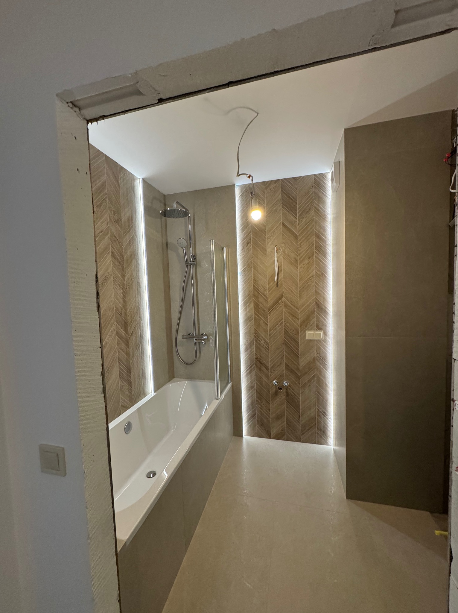 Nowoczesna łazienka z wanną, szklanym parawanem i oświetleniem LED wzdłuż ściany z wzorem jodełki. Widoczny fragment surowego otworu drzwiowego.