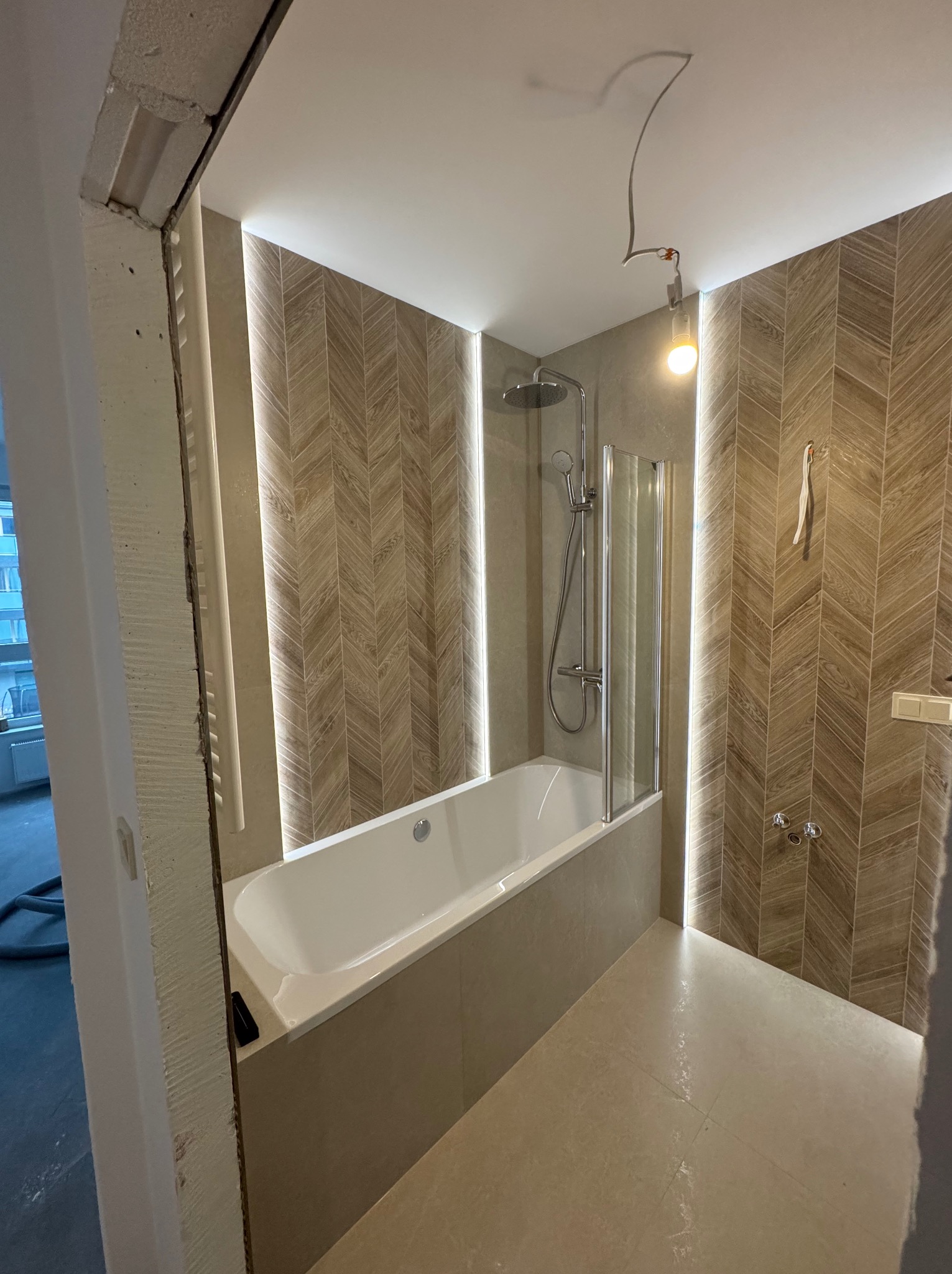 Nowoczesna łazienka z wanną, kabiną prysznicową i podświetlanymi płytkami w jodełkę. Widoczne niedokończone wykończenia przy futrynie drzwi.