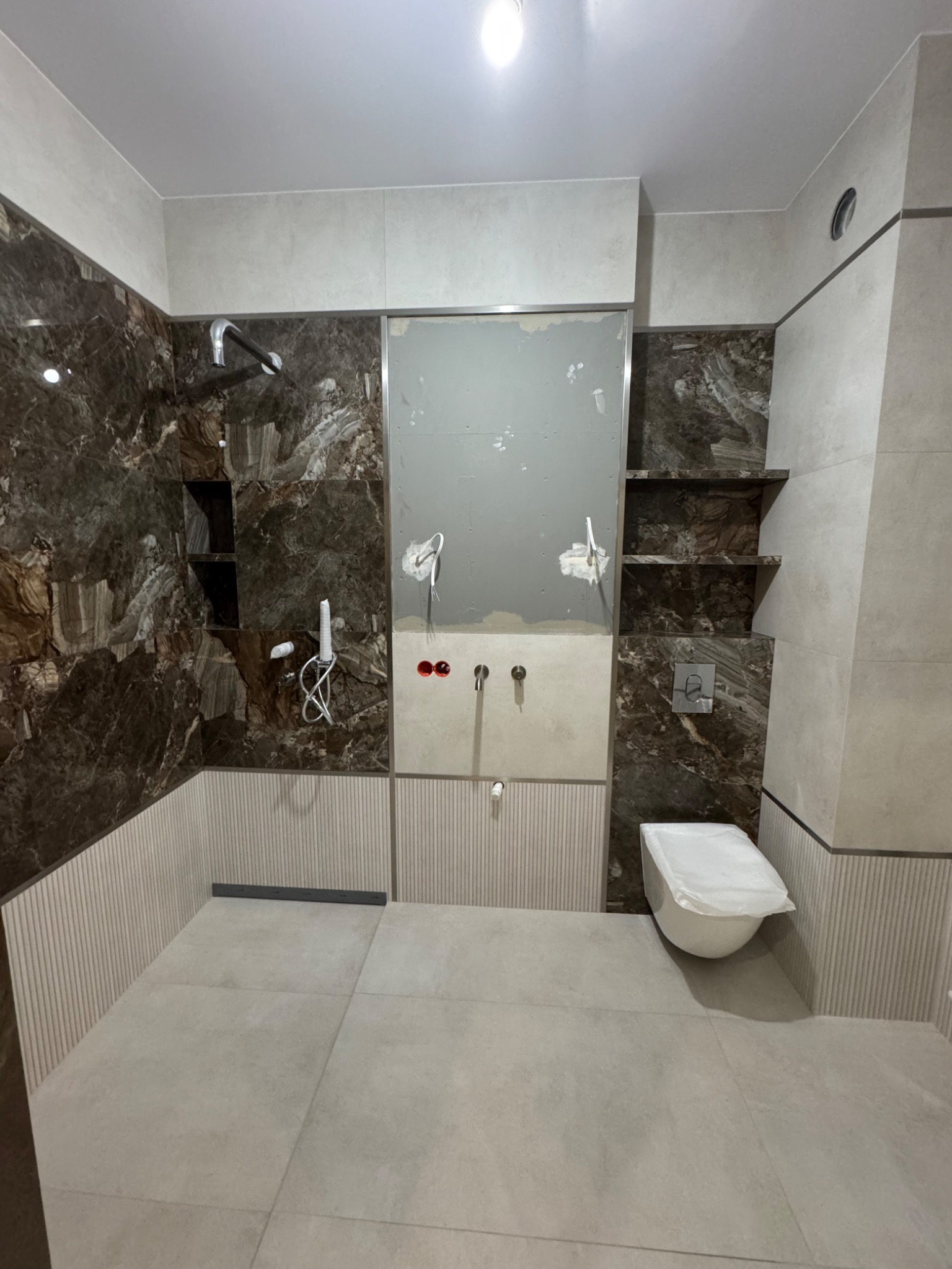 Łazienka w trakcie wykończenia: brązowe płytki imitujące marmur w strefie prysznica z półkami, częściowo zamontowana armatura, zabezpieczona toaleta, jasne płytki na podłodze.