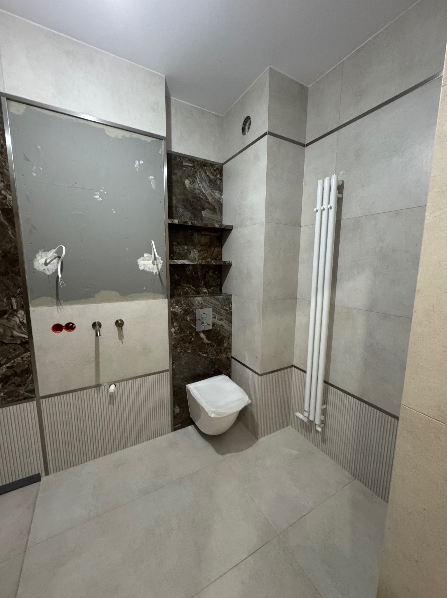 Łazienka w trakcie wykańczania: płytki w odcieniach szarości i beżu, zabudowa z półkami z ciemnego marmuru, biały grzejnik. Widoczne przygotowane instalacje wodne.