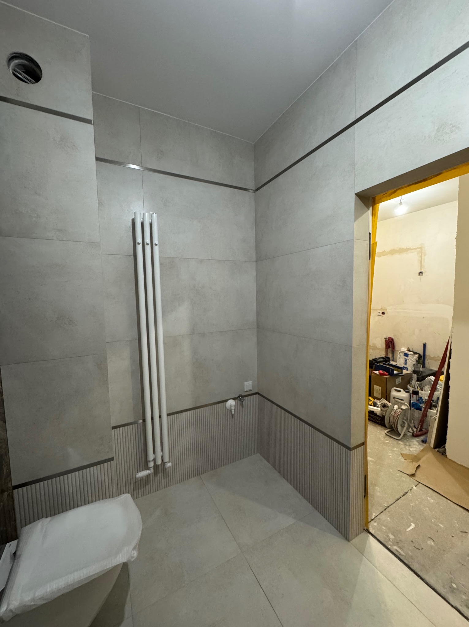 Jasna łazienka po remoncie z grzejnikiem drabinkowym, wykończona płytkami w odcieniach szarości z dekoracyjnym pasem. W tle widoczne narzędzia i materiały budowlane.