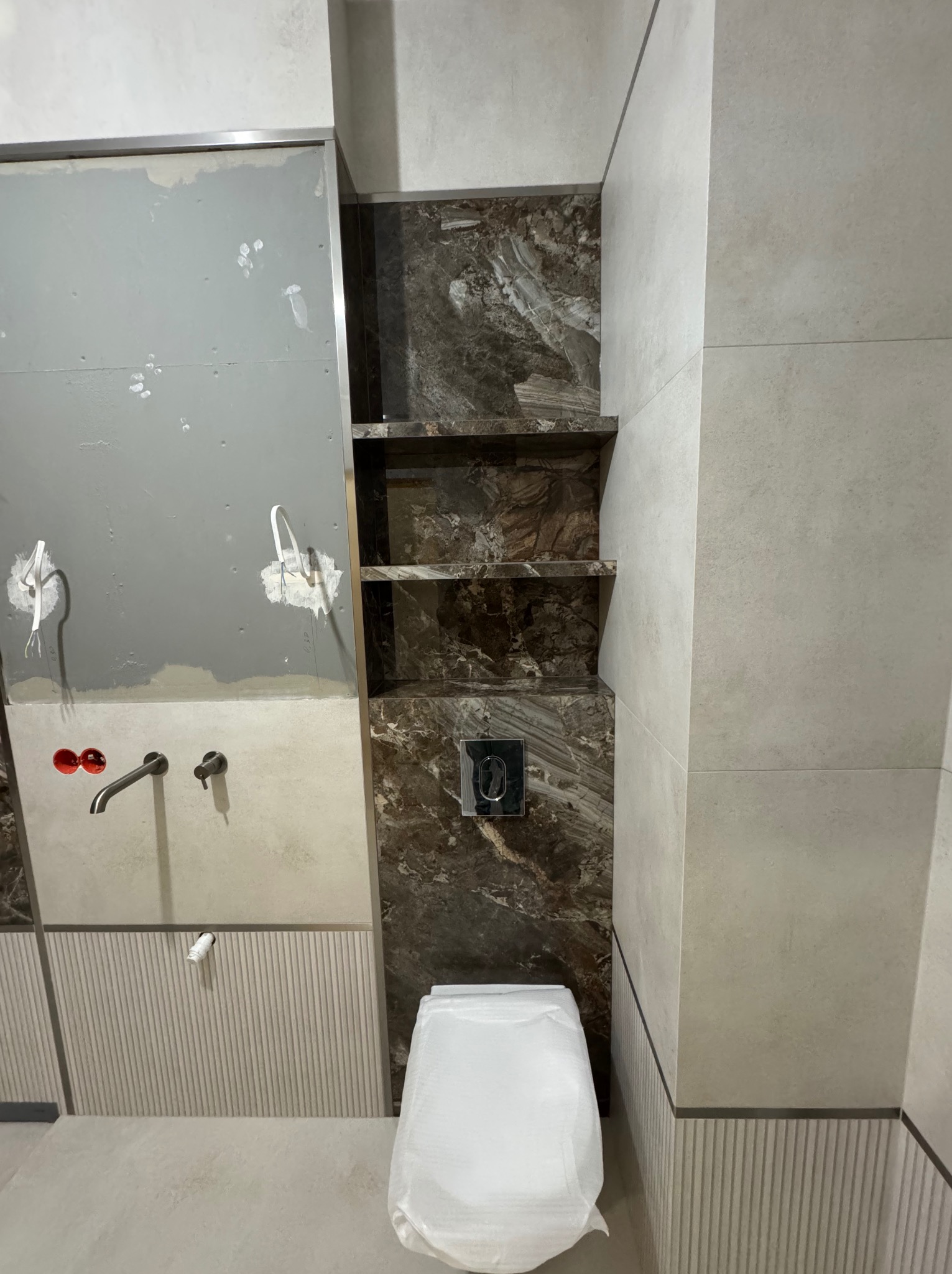 Łazienka w trakcie wykańczania: widoczne płytki imitujące marmur, półki wnękowe, armatura i częściowo wykończona ściana z instalacją. Toaleta zabezpieczona folią.