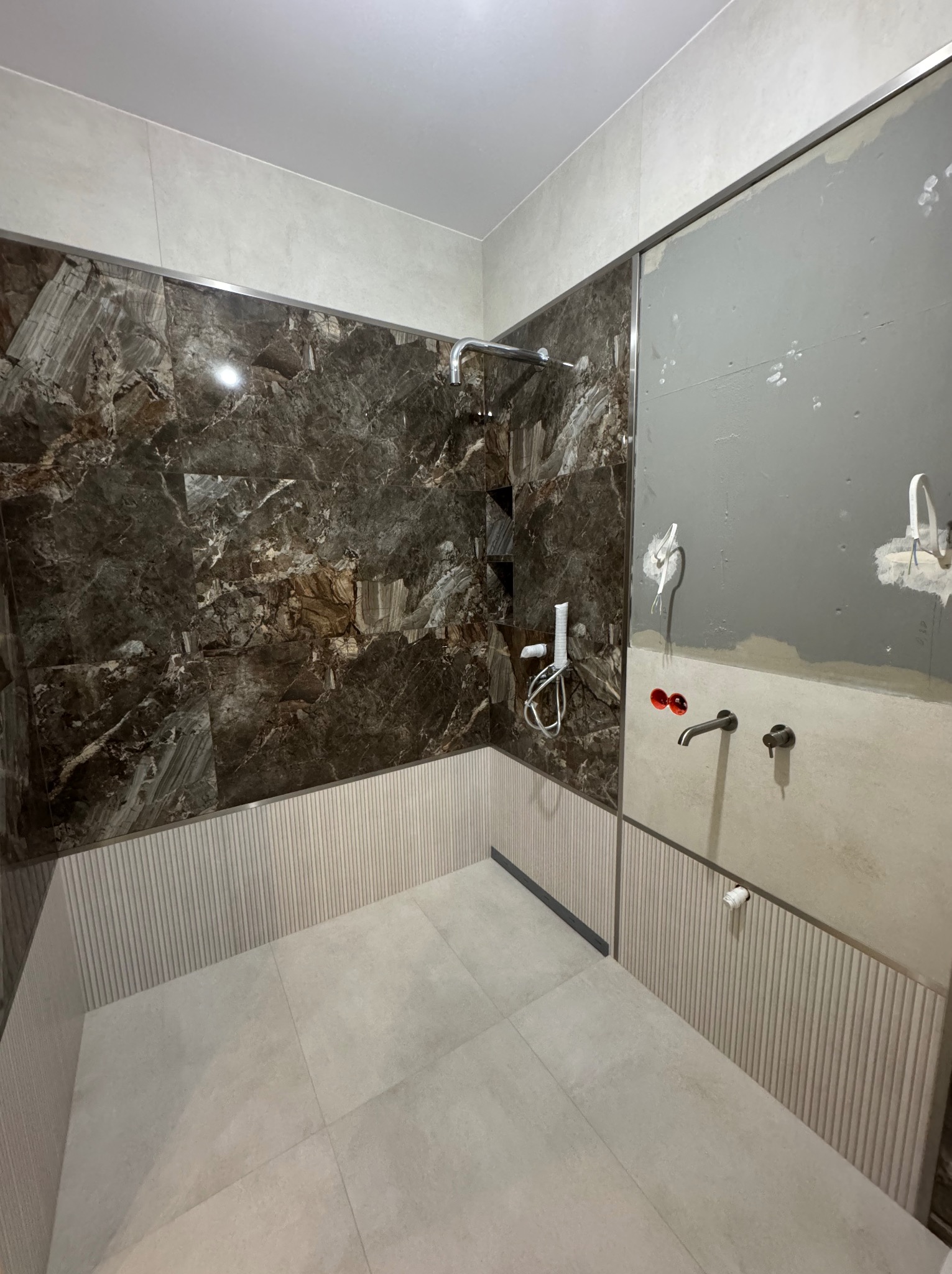 Nowoczesna łazienka w trakcie wykańczania: marmurowe płytki, armatura w kolorze czarnym, surowy beton na ścianie. Widoczne elementy instalacji wodnej.