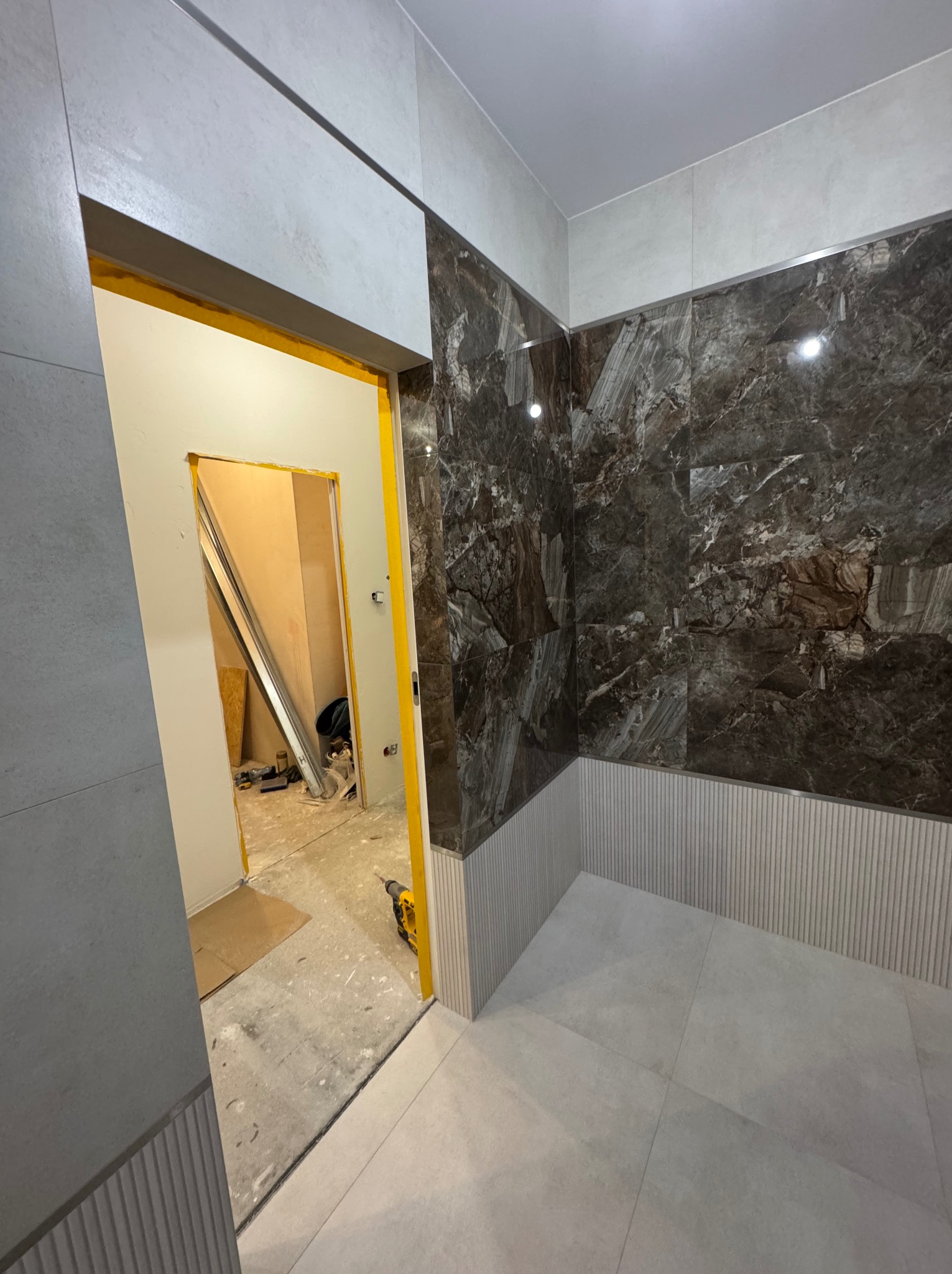 Częściowo wykończona łazienka z marmurowymi płytkami na ścianach i jasnymi płytkami na podłodze. Widoczne narzędzia i materiały budowlane w sąsiednim pomieszczeniu.