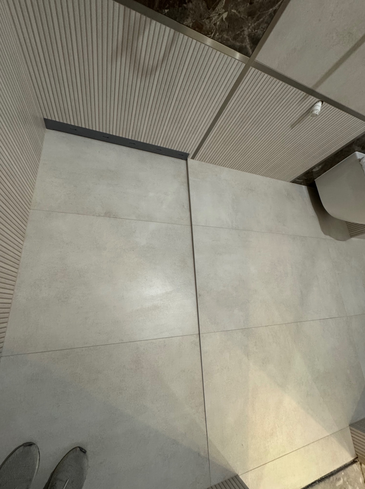 Wykończona łazienka z beżowymi płytkami na ścianach i podłodze, widoczny fragment sufitu z oświetleniem. Minimalistyczny design z ukrytym odpływem liniowym.