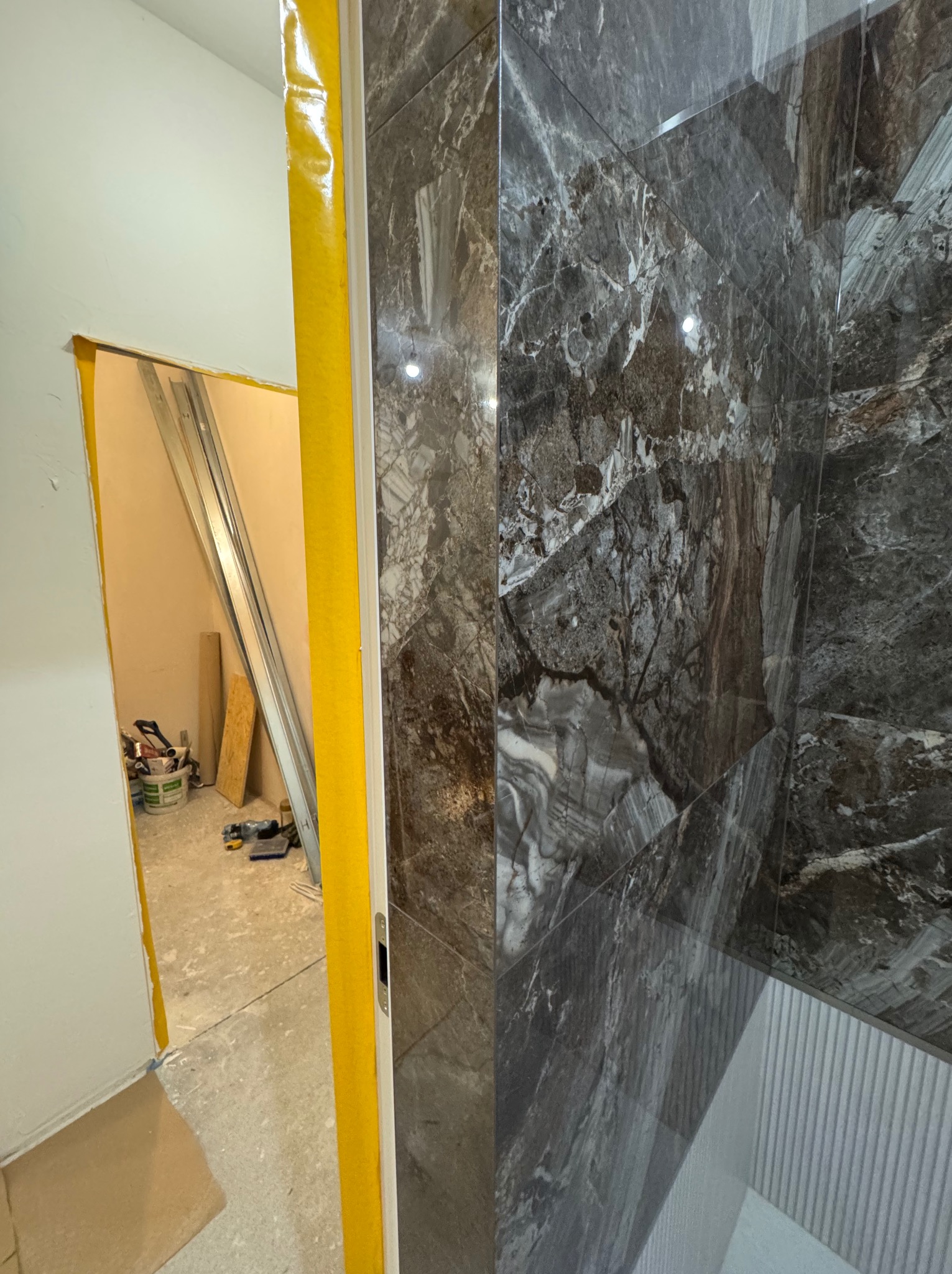 Fragment łazienki w trakcie wykończenia: ciemne płytki imitujące marmur na ścianie, widoczne narzędzia i profile w przejściu drzwiowym zabezpieczonym żółtą taśmą.