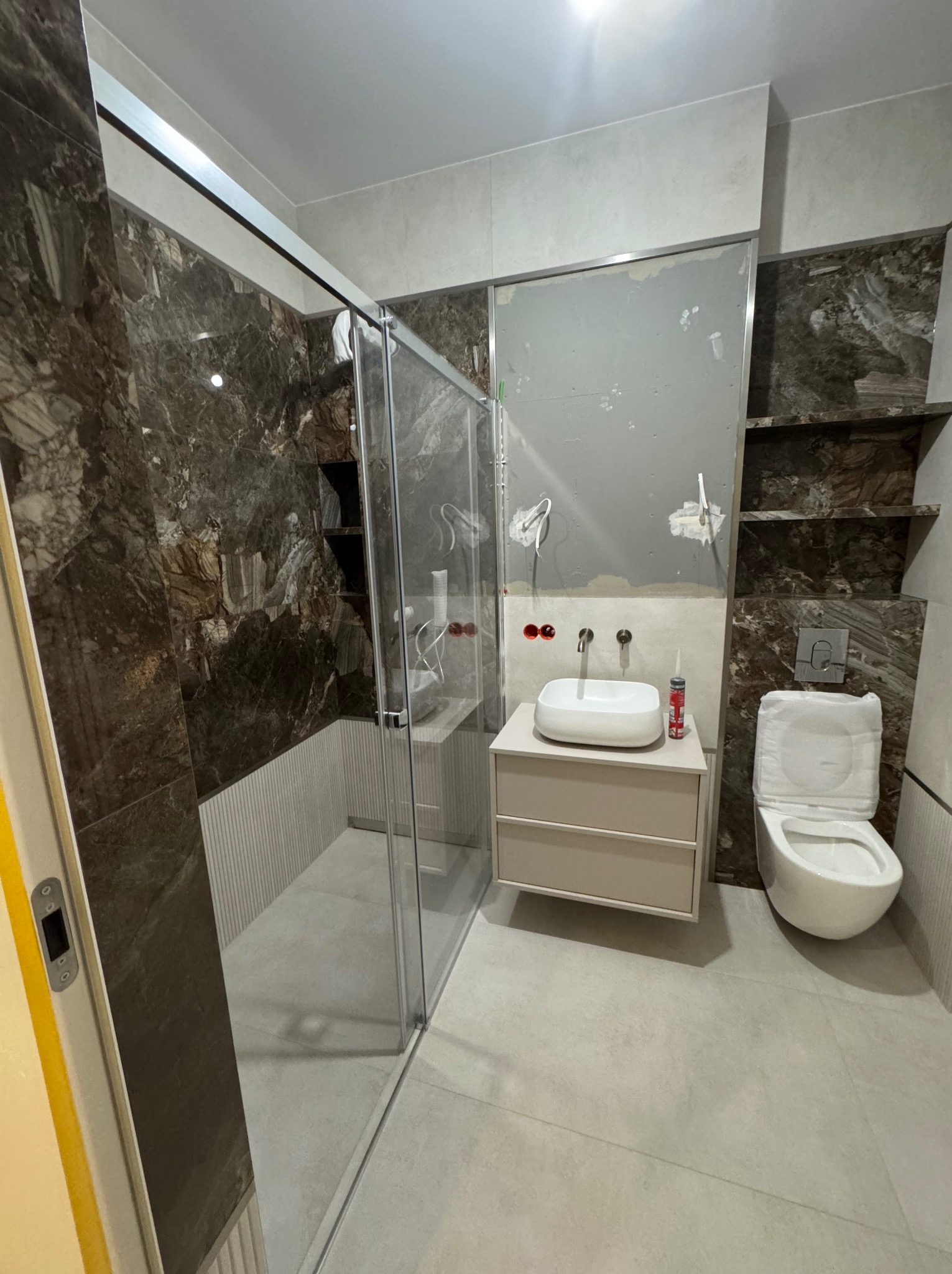 Nowoczesna łazienka w trakcie wykańczania: kabina prysznicowa ze szklanymi drzwiami, umywalka na szafce, toaleta i marmurowe płytki z półkami wnękowymi.