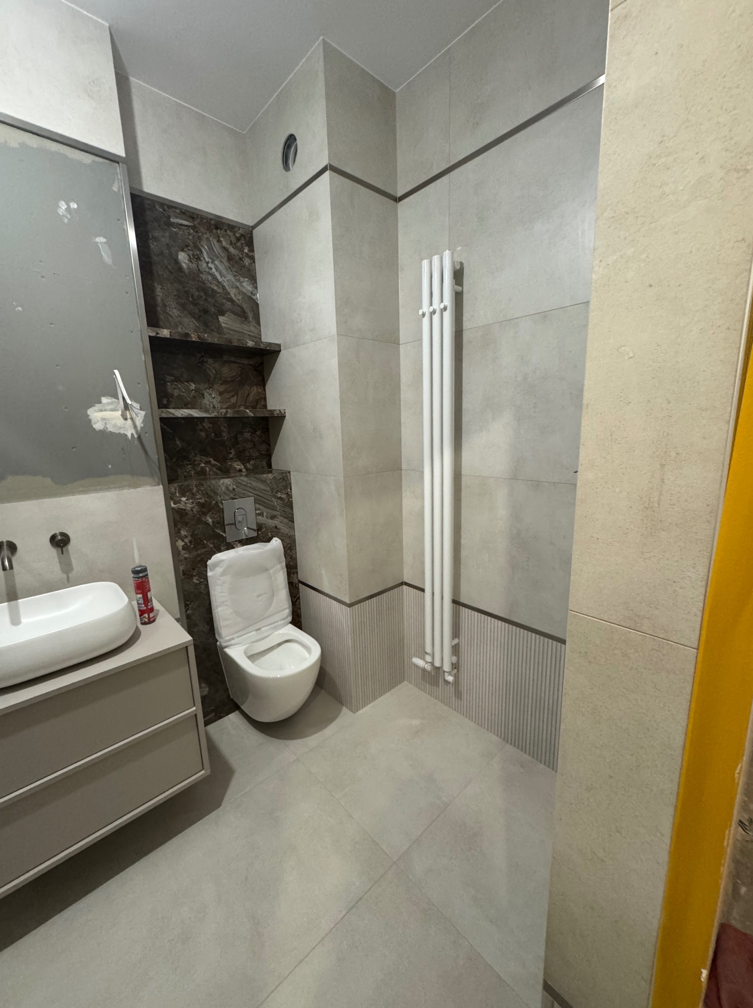 Jasna łazienka po remoncie: umywalka z szafką, toaleta, grzejnik drabinkowy, półki z ciemnego marmuru. Widoczne ślady po remoncie na ścianie.