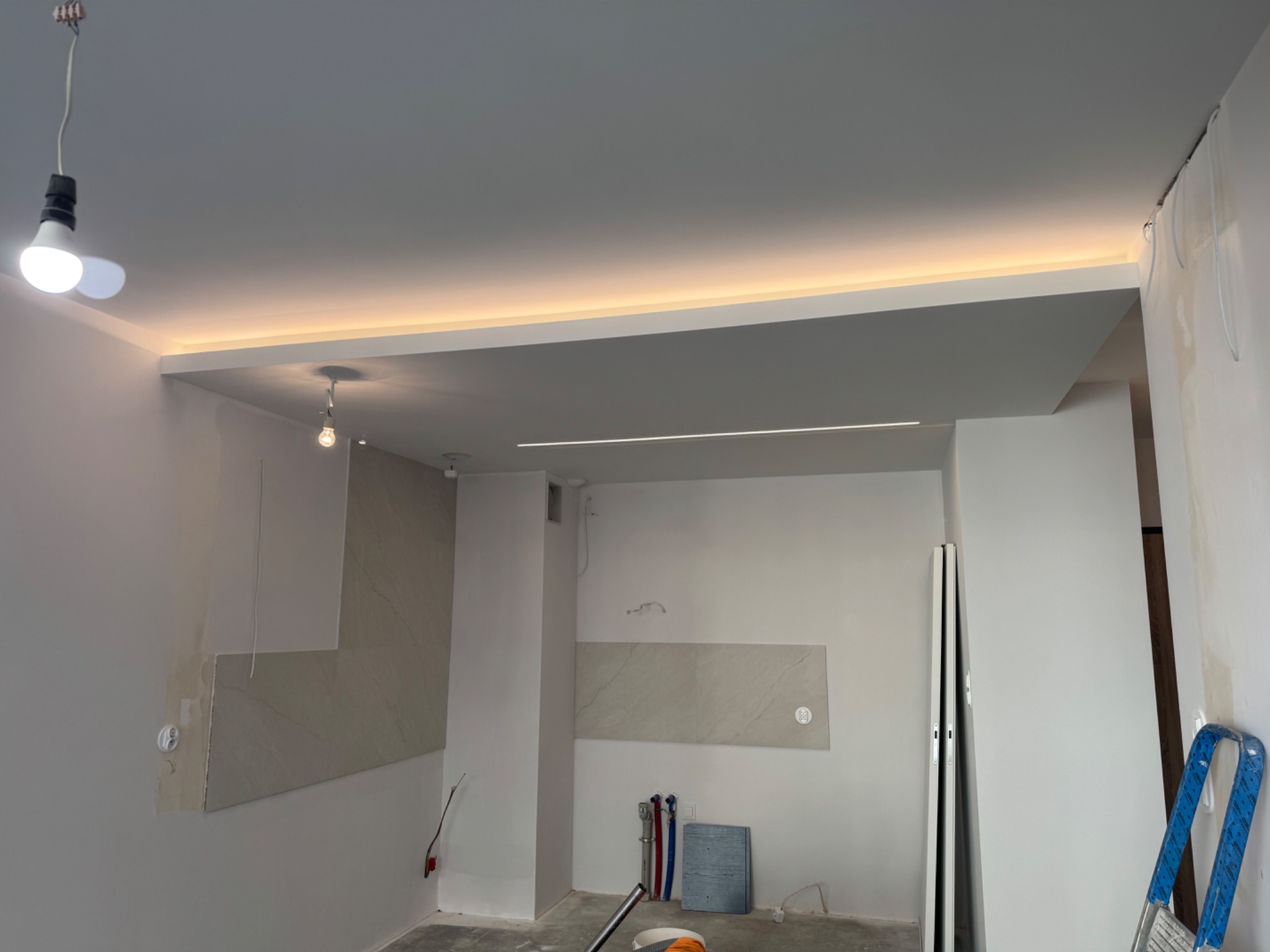 Wnętrze w trakcie wykańczania: częściowo położone płytki, podwieszany sufit z oświetleniem LED, widoczne instalacje i drabina. W tle surowy beton i wisząca żarówka.
