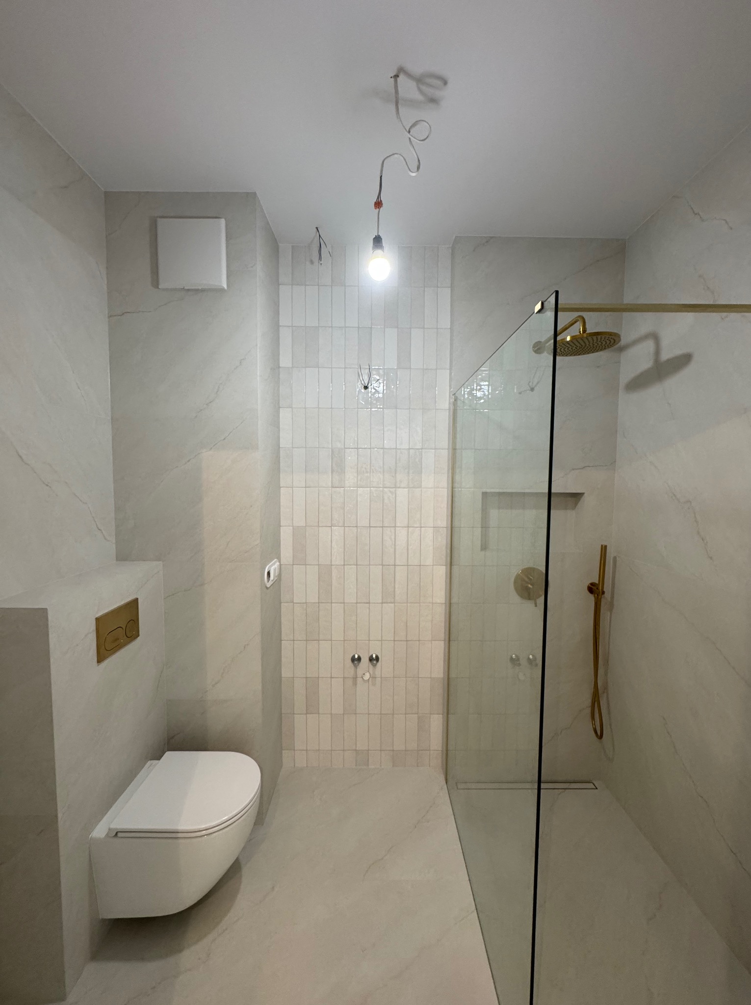 Nowoczesna łazienka z kabiną prysznicową typu walk-in, wykończona płytkami imitującymi marmur i złotą armaturą. Widoczne podwieszane WC i minimalistyczny design.