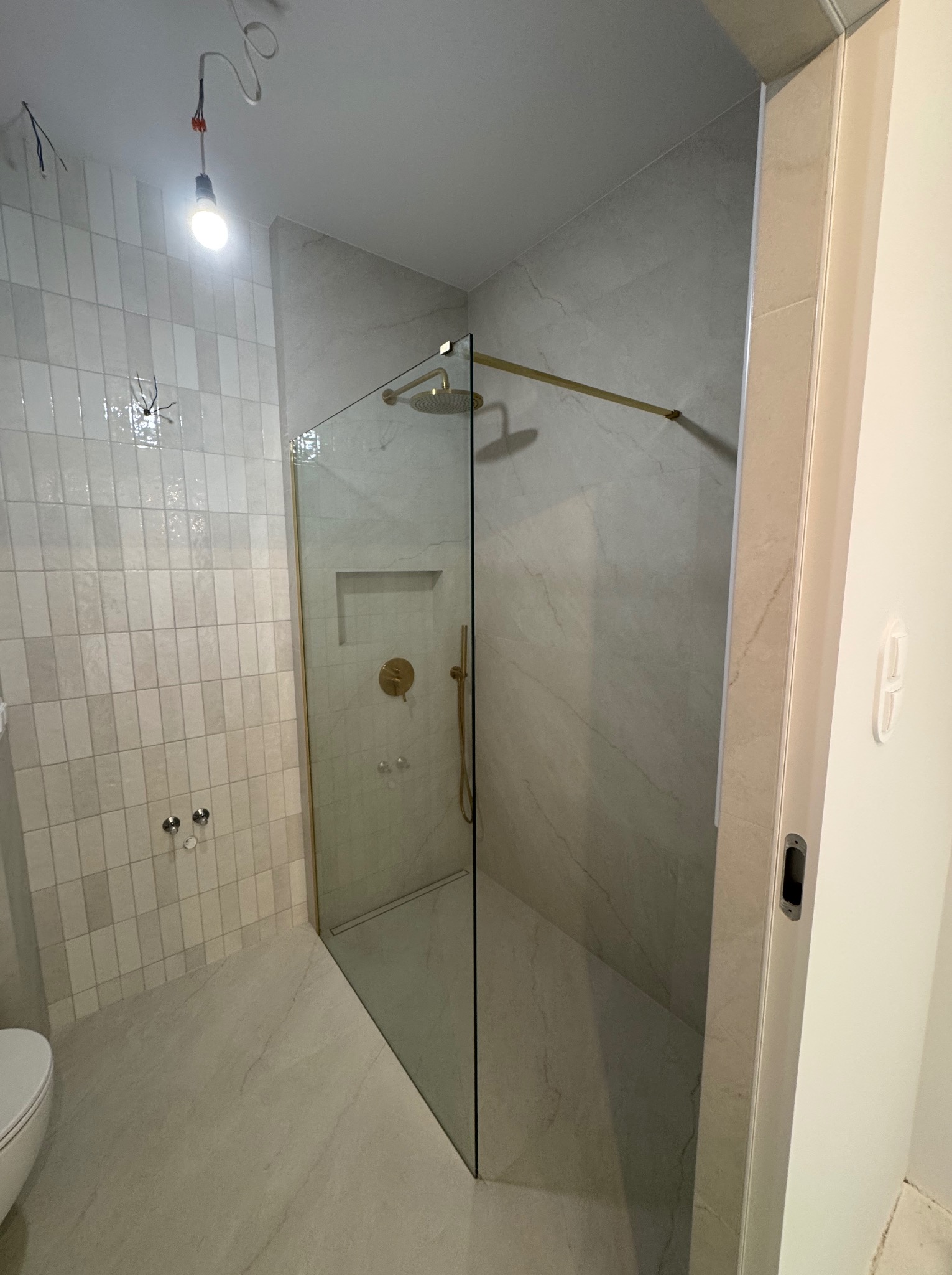 Wykończona łazienka z kabiną prysznicową typu walk-in, złotymi elementami armatury i białymi, pionowo ułożonymi płytkami. Widoczne niedokończone podłączenie oświetlenia.