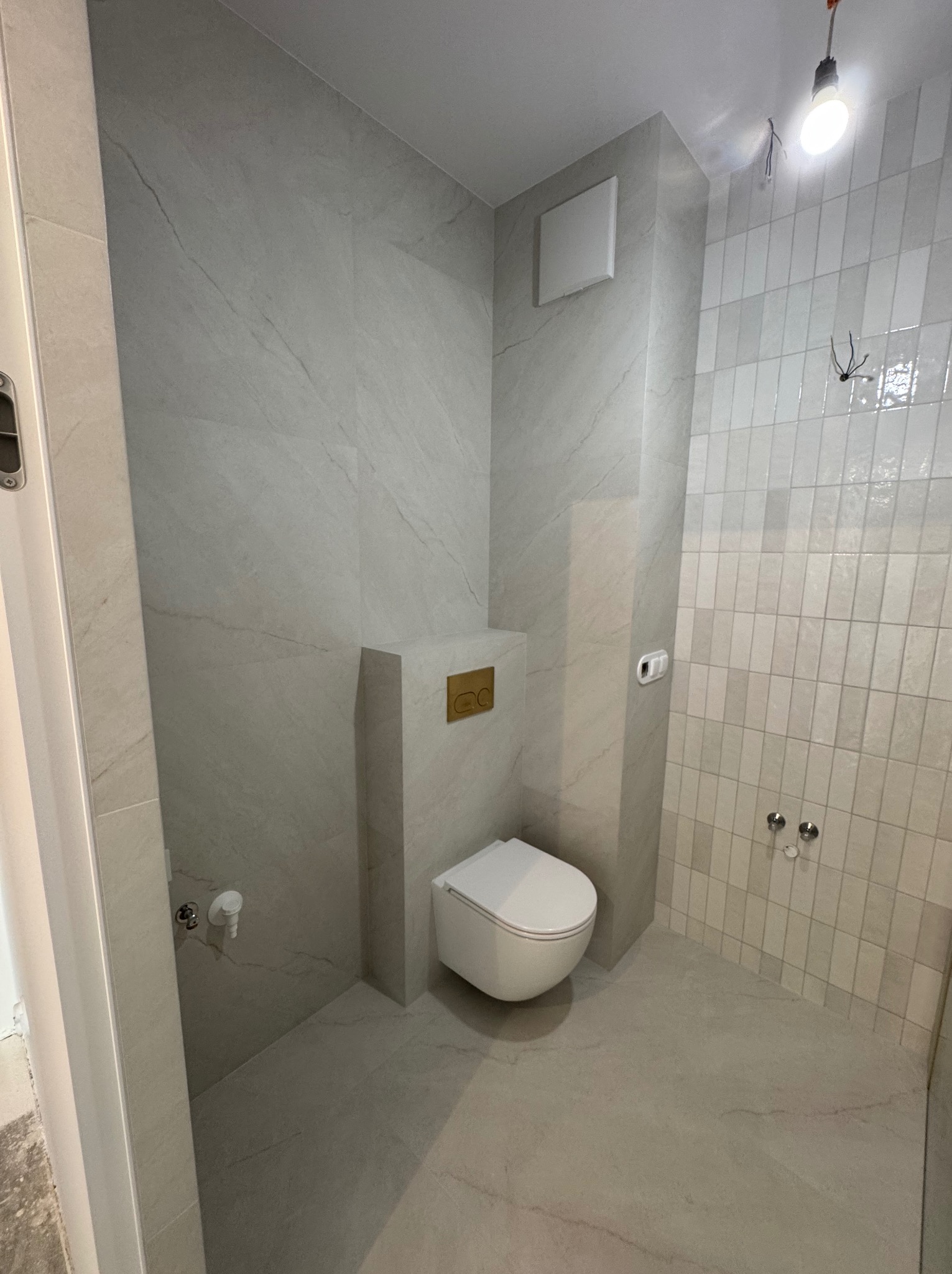 Nowoczesna łazienka w odcieniach szarości z podwieszaną toaletą i płytkami imitującymi marmur. Widoczne przyłącza elektryczne i wentylacja.