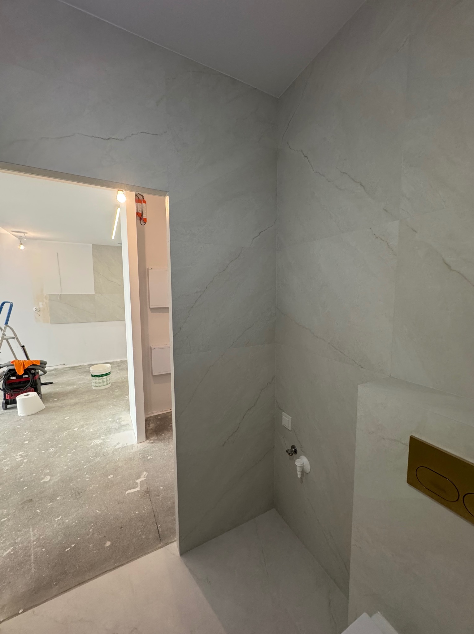 Wykończona ściana łazienki z szarymi płytkami imitującymi marmur, widoczny fragment toalety i instalacja wodna. W tle pomieszczenie w trakcie remontu.