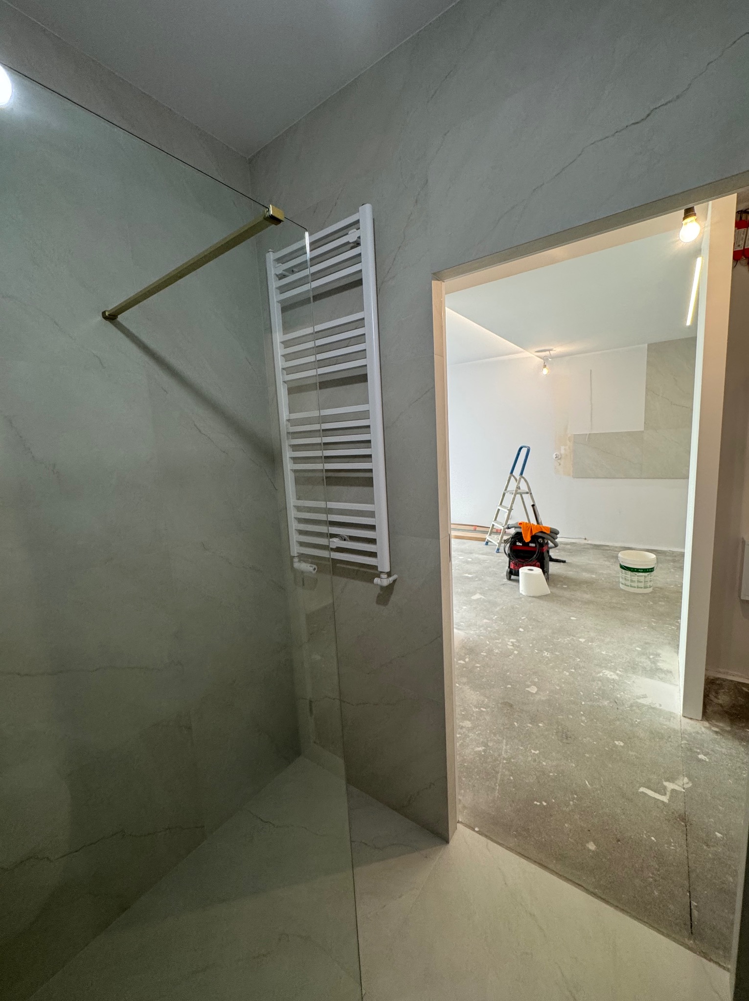 Nowoczesna łazienka w trakcie wykończenia: szklana kabina z złotymi elementami, biały grzejnik drabinkowy, widok na remontowane wnętrze z drabiną i narzędziami.