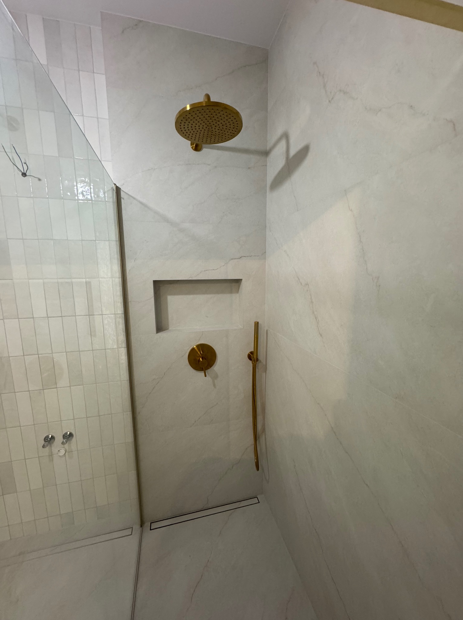 Nowoczesna kabina prysznicowa z białymi płytkami imitującymi marmur i złotą armaturą. Widoczny odpływ liniowy i wnęka w ścianie.