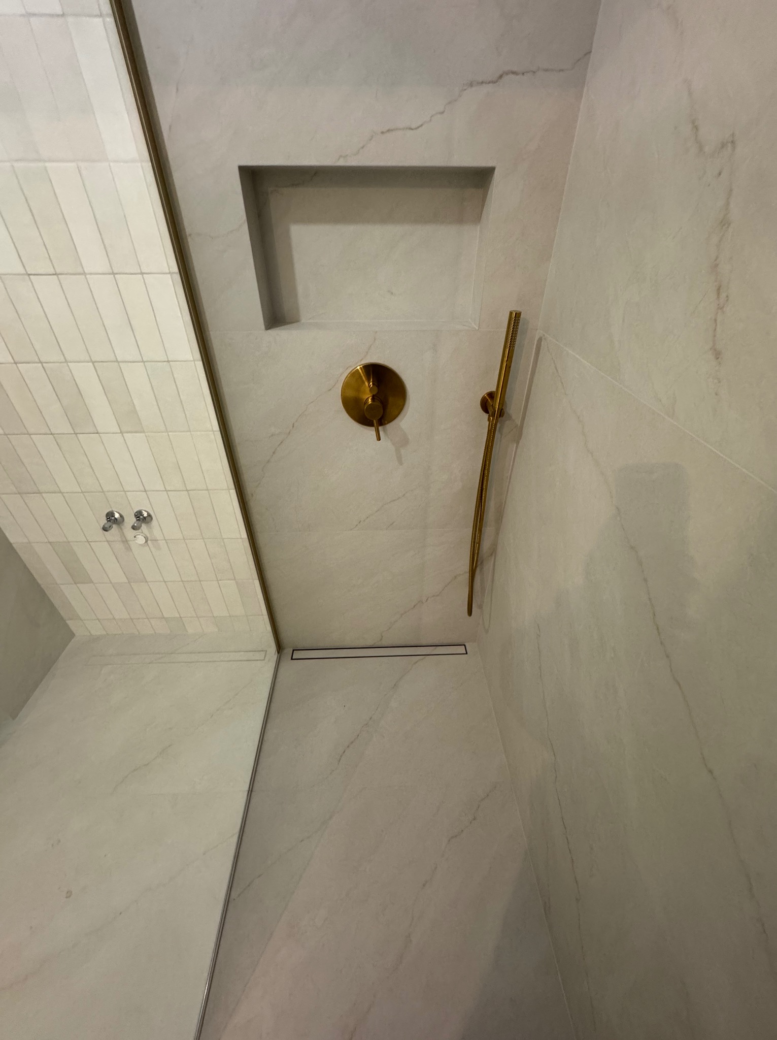 Nowoczesna kabina prysznicowa z marmurowymi płytkami, złotą armaturą i wnęką. Minimalistyczny design z odpływem liniowym i subtelnym oświetleniem.