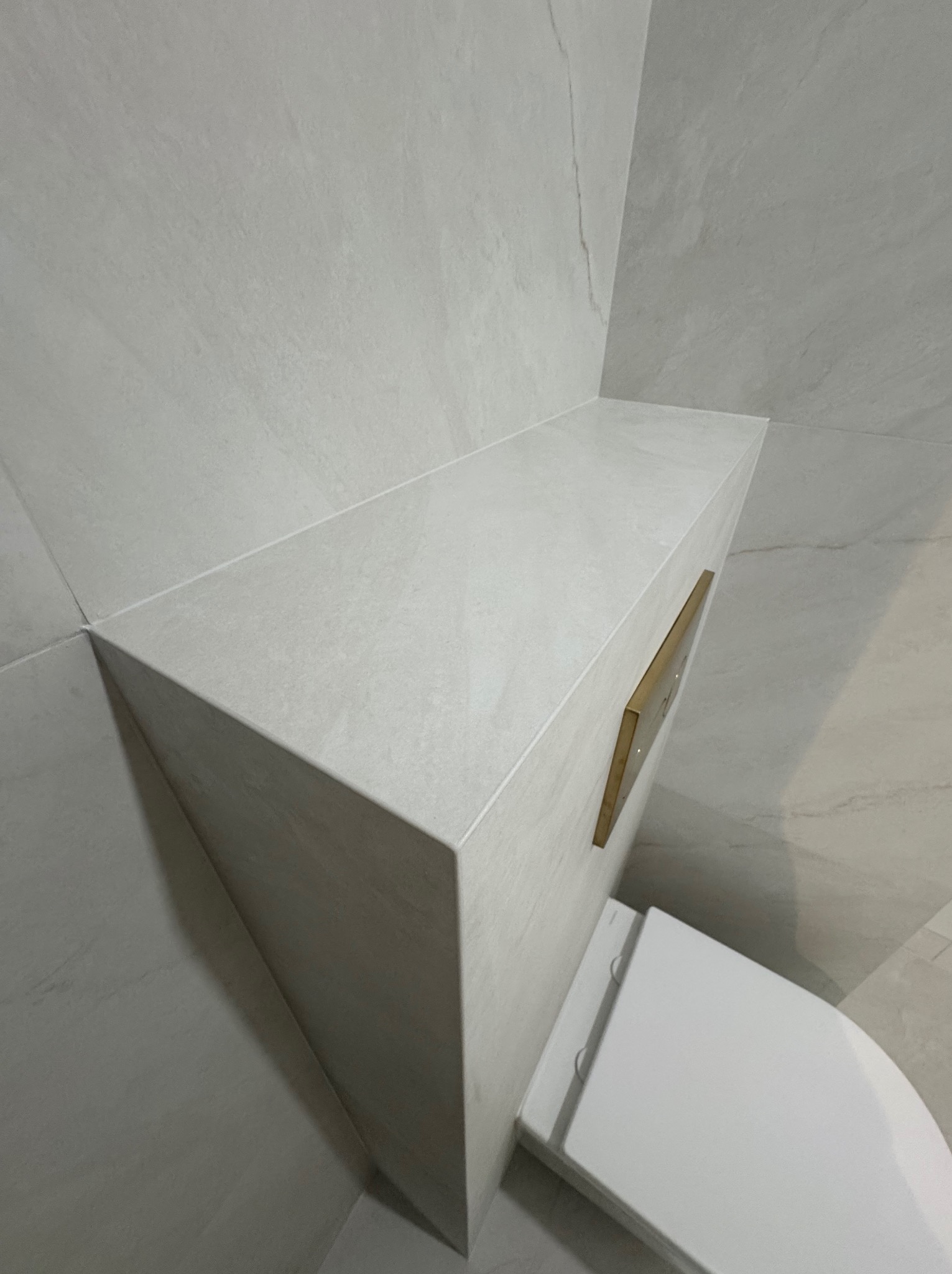 Minimalistyczna łazienka: zabudowa WC z szarej, matowej płytki imitującej kamień, zlicowana ze ścianą, z złotym przyciskiem spłukującym i białą miską ustępową.