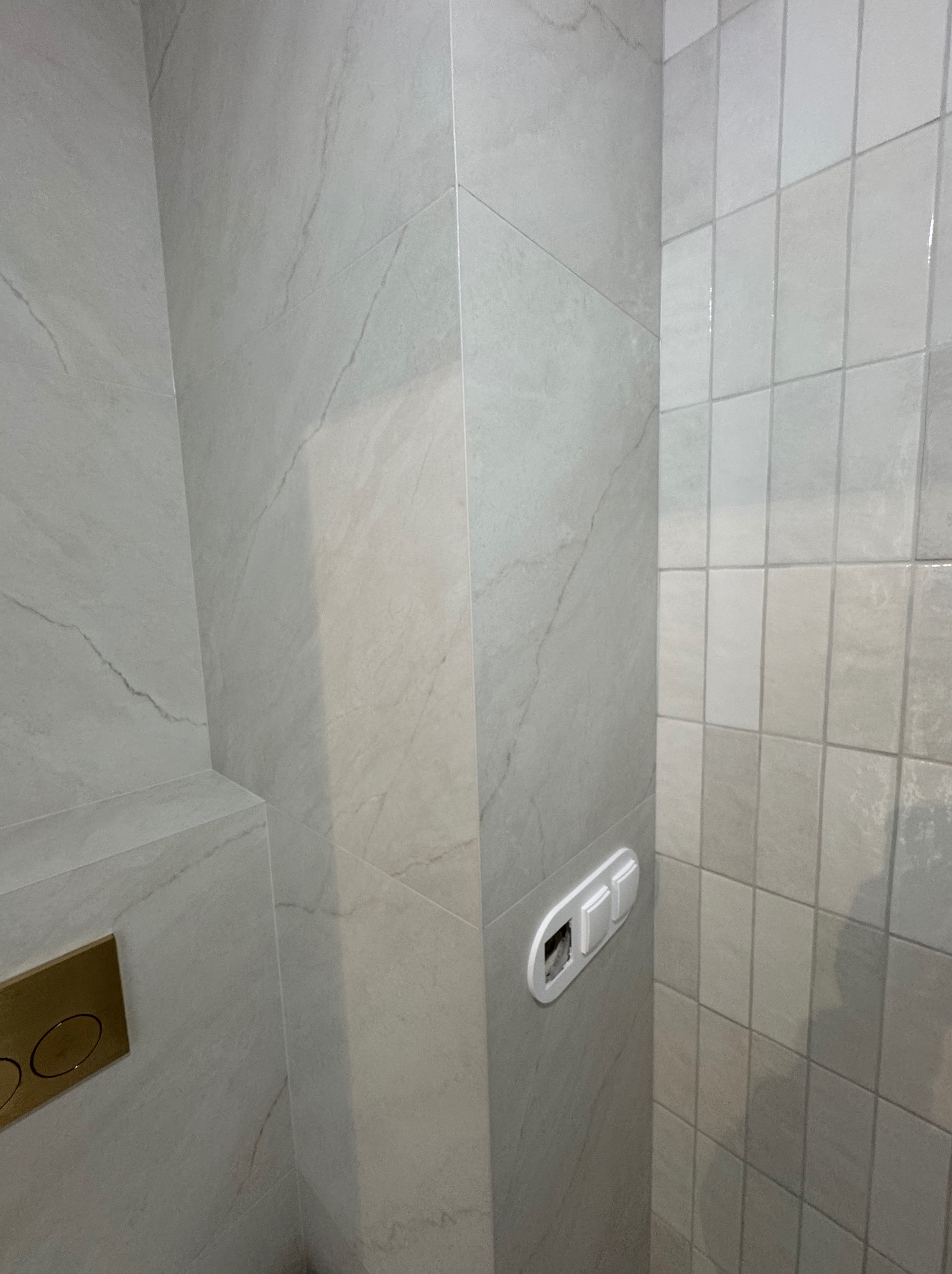 Wykończona łazienka z szarymi płytkami imitującymi marmur, widoczny panel WC w kolorze złotym i włączniki światła. Minimalistyczny design.