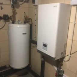 Biały zasobnik ciepłej wody z manometrem i biała jednostka grzewcza Aquami Otensa z wyświetlaczem, połączone rurami w pomieszczeniu z beżowymi płytkami.