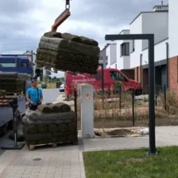 Rozładunek palet z trawą rolowaną za pomocą dźwigu samochodowego na tle nowoczesnych domów jednorodzinnych, widoczny pracownik w niebieskiej koszulce.