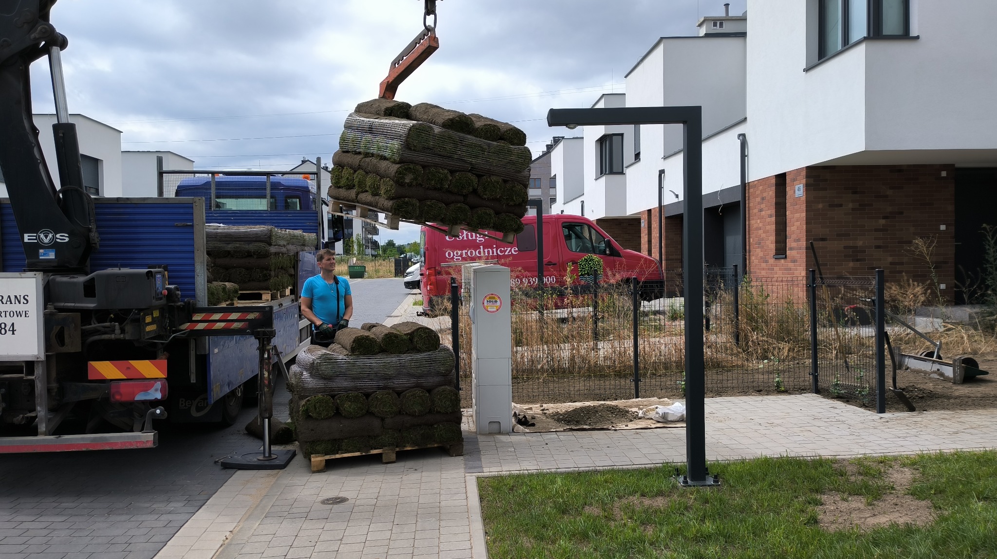 Rozładunek palet z trawą rolowaną za pomocą dźwigu samochodowego na tle nowoczesnych domów jednorodzinnych, widoczny pracownik w niebieskiej koszulce.