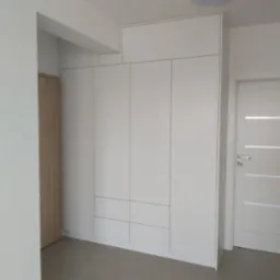 Biała szafa wnękowa z frontami bez uchwytów, dopasowana do wysokości pomieszczenia, z widocznymi trzema szufladami w dolnej części, obok białe drzwi z poziomymi przeszkleniami.