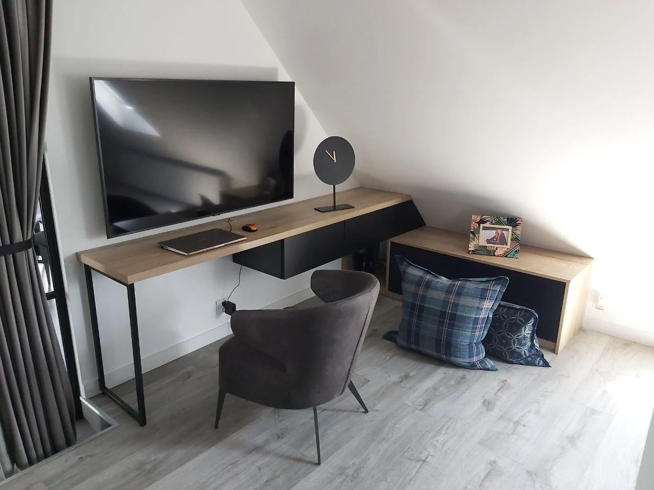 Nowoczesny pokój na poddaszu z biurkiem z drewnianym blatem na metalowych nogach, szafką wiszącą z czarnymi frontami, telewizorem na ścianie, szarą tapicerowaną krzesłem i elementami dekoracyjnymi.