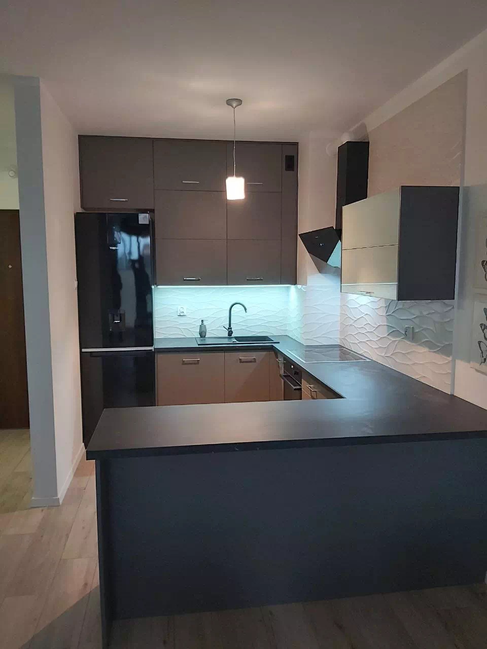 Nowoczesna kuchnia z ciemnymi górnymi szafkami, jasnymi dolnymi i czarnym blatem, oświetleniem LED pod szafkami i dekoracyjnymi białymi płytkami na ścianie.