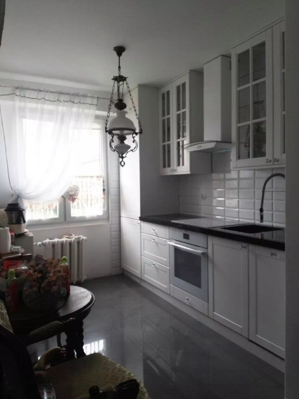 Jasna kuchnia z białymi szafkami z przeszklonymi frontami, czarnym blatem i wiszącą lampą w stylu retro, widok z perspektywy jadalni.