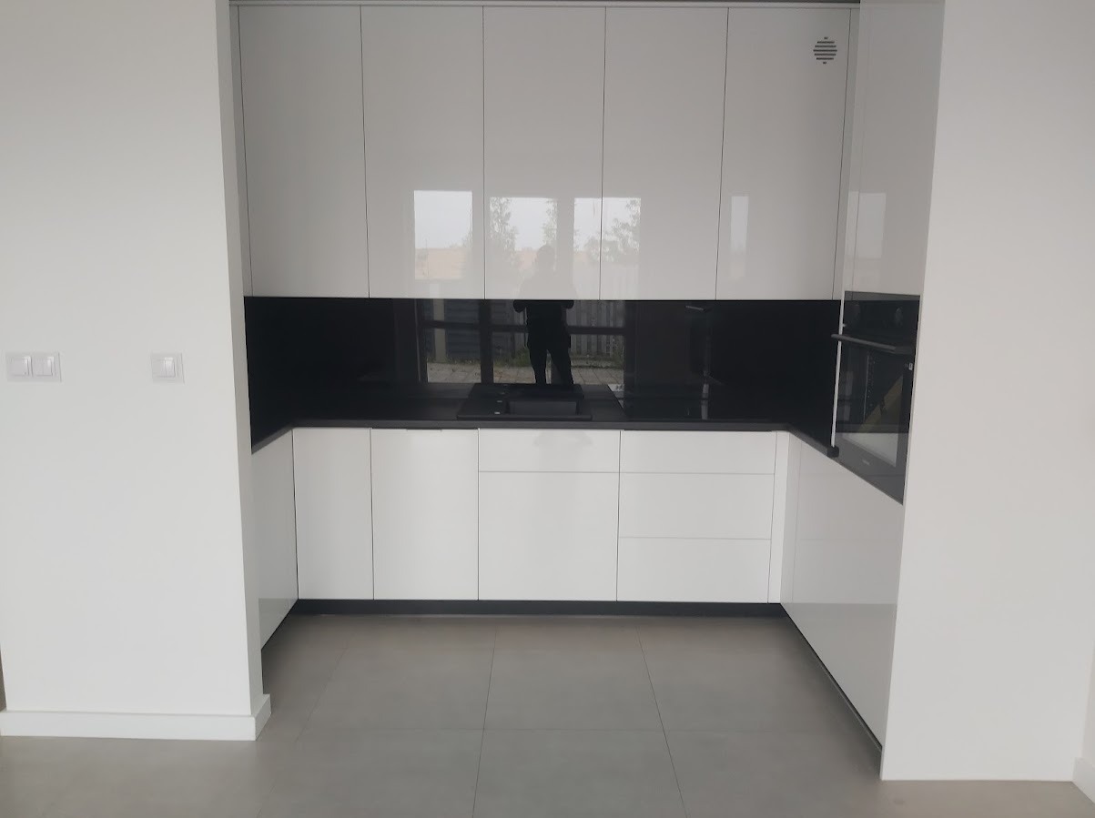 Biała kuchnia z połyskiem, czarny panel nad blatem, minimalistyczny design, widoczne odbicie osoby w panelu.