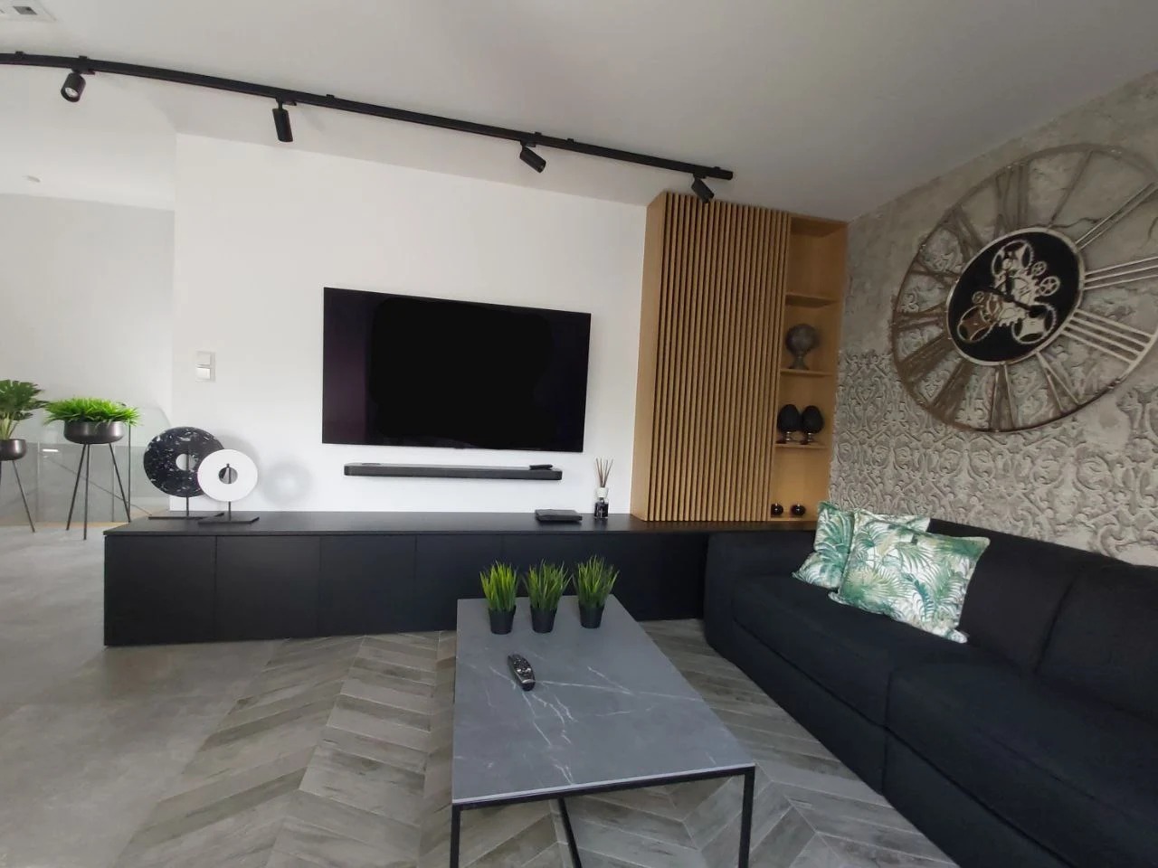 Nowoczesny salon z czarną szafką RTV, drewnianą ścianką działową, szarą podłogą w jodełkę i dekoracyjnym zegarem na ścianie.