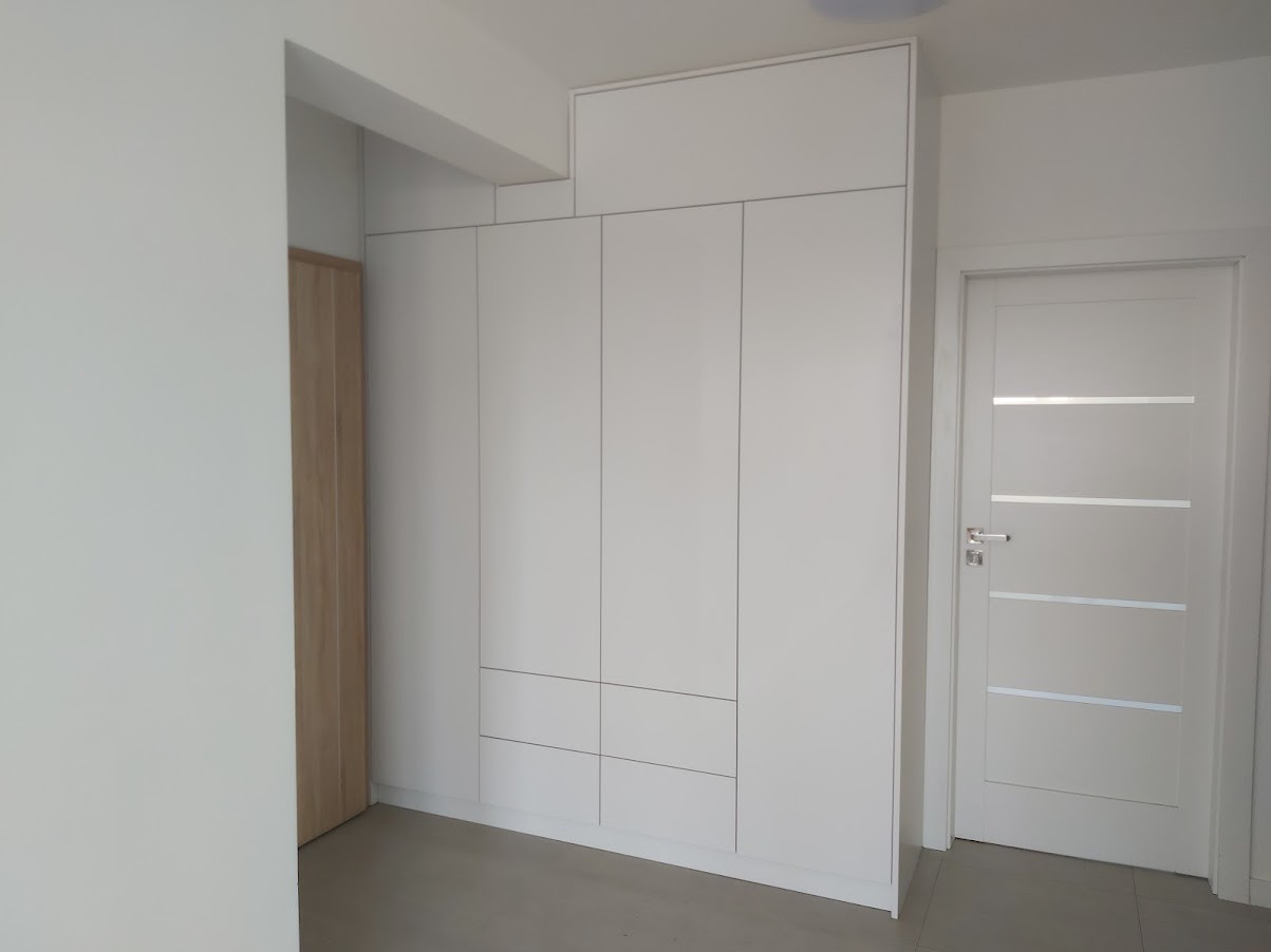 Biała szafa wnękowa z frontami bez uchwytów, dopasowana do wysokości pomieszczenia, z widocznymi trzema szufladami w dolnej części, obok białe drzwi z poziomymi przeszkleniami.