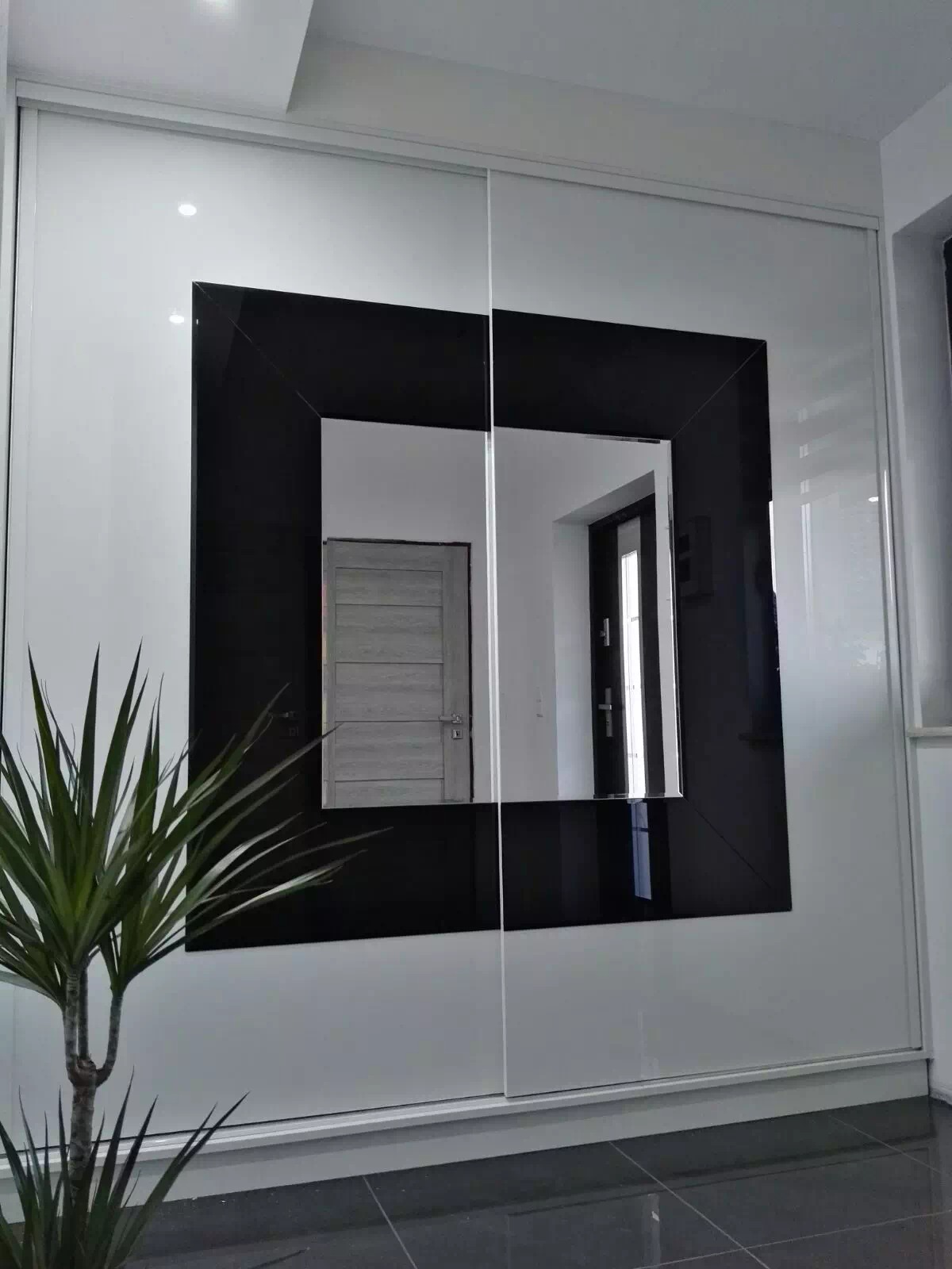 Biała szafa przesuwna z czarną, kwadratową ramą lustra, odbijająca wnętrze pokoju i drzwi, z rośliną doniczkową w lewym dolnym rogu.