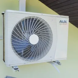 Zewnętrzna jednostka klimatyzacji AUX DC Inverter zamontowana na żółtej ścianie budynku, widoczna z boku, z fragmentem brązowego dachu w tle.