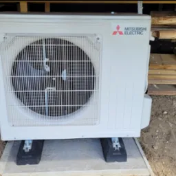 Zewnętrzna jednostka Mitsubishi Electric, zamontowana na betonowej podstawie z gumowymi podkładkami, widoczna na tle ziemi i elementów konstrukcyjnych budynku.