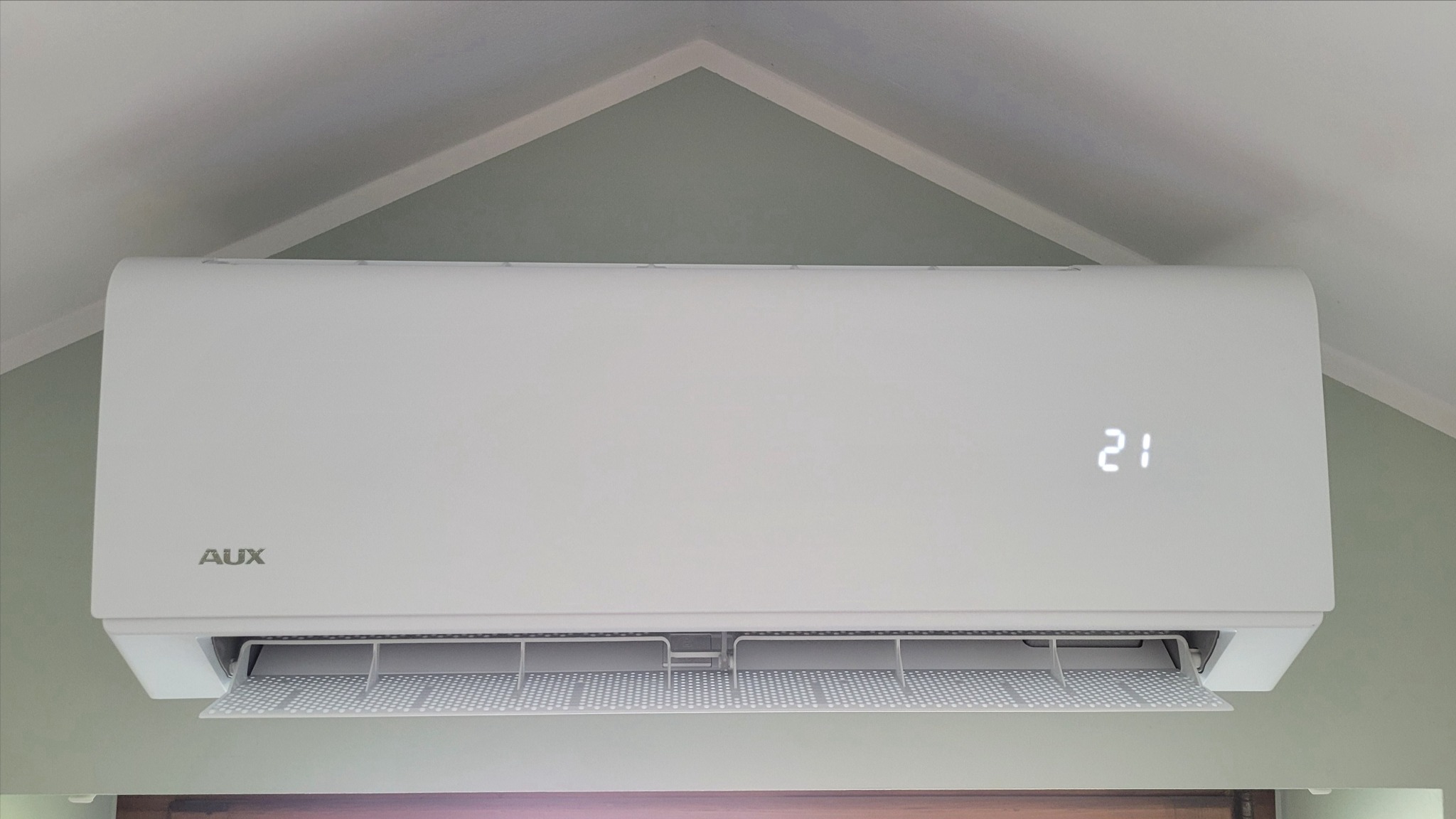 Biały klimatyzator marki AUX zamontowany na ścianie, wyświetlający temperaturę 21 stopni Celsjusza, z widocznym filtrem powietrza.