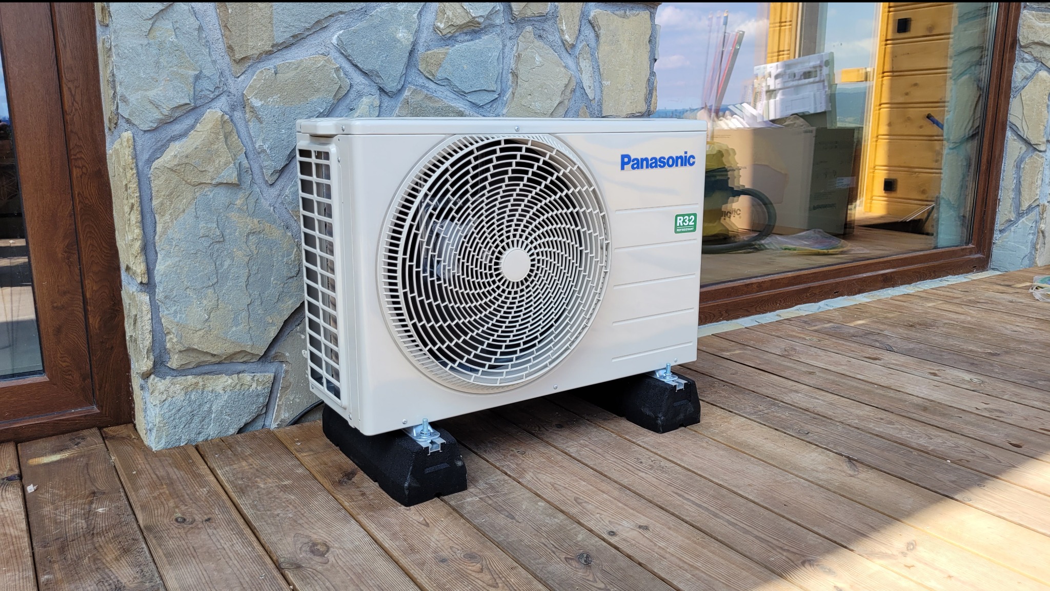 Zewnętrzna jednostka klimatyzatora Panasonic R32 zamontowana na czarnych podkładkach antywibracyjnych na drewnianym tarasie, widoczna ściana wyłożona kamieniem i okno z brązową ramą.