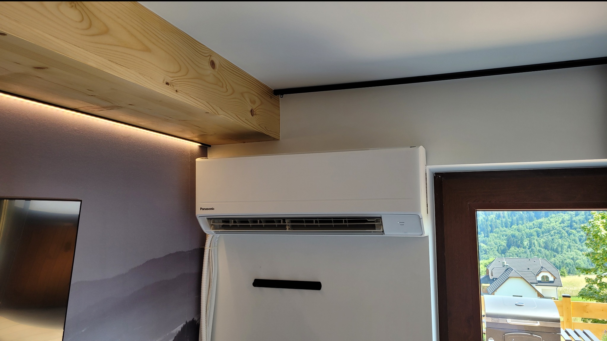 Biała jednostka wewnętrzna klimatyzacji Panasonic na ścianie z tapetą górską, widok przez okno na zalesione wzgórza, drewniana belka stropowa z oświetleniem LED.