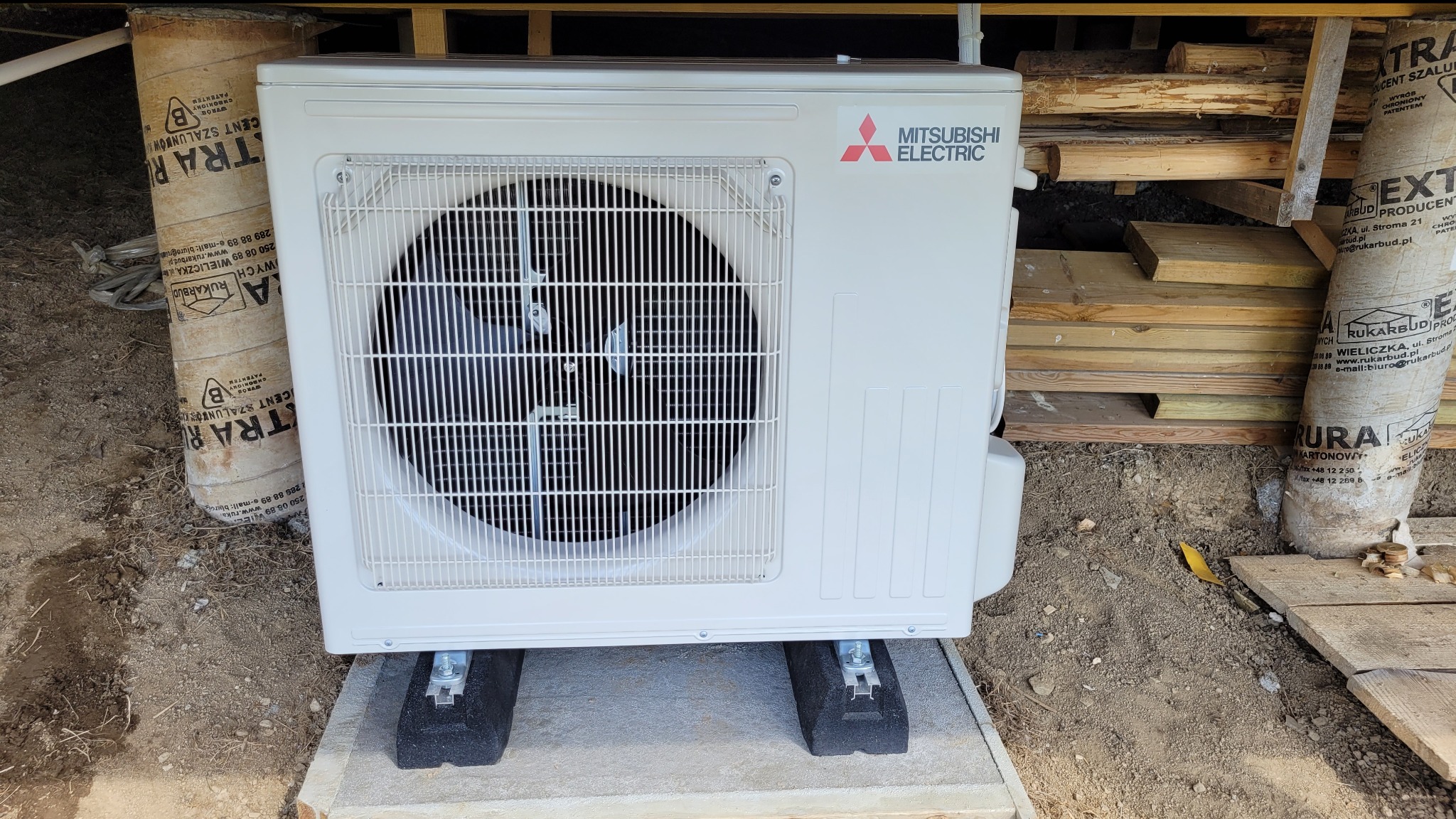 Zewnętrzna jednostka Mitsubishi Electric, zamontowana na betonowej podstawie z gumowymi podkładkami, widoczna na tle ziemi i elementów konstrukcyjnych budynku.
