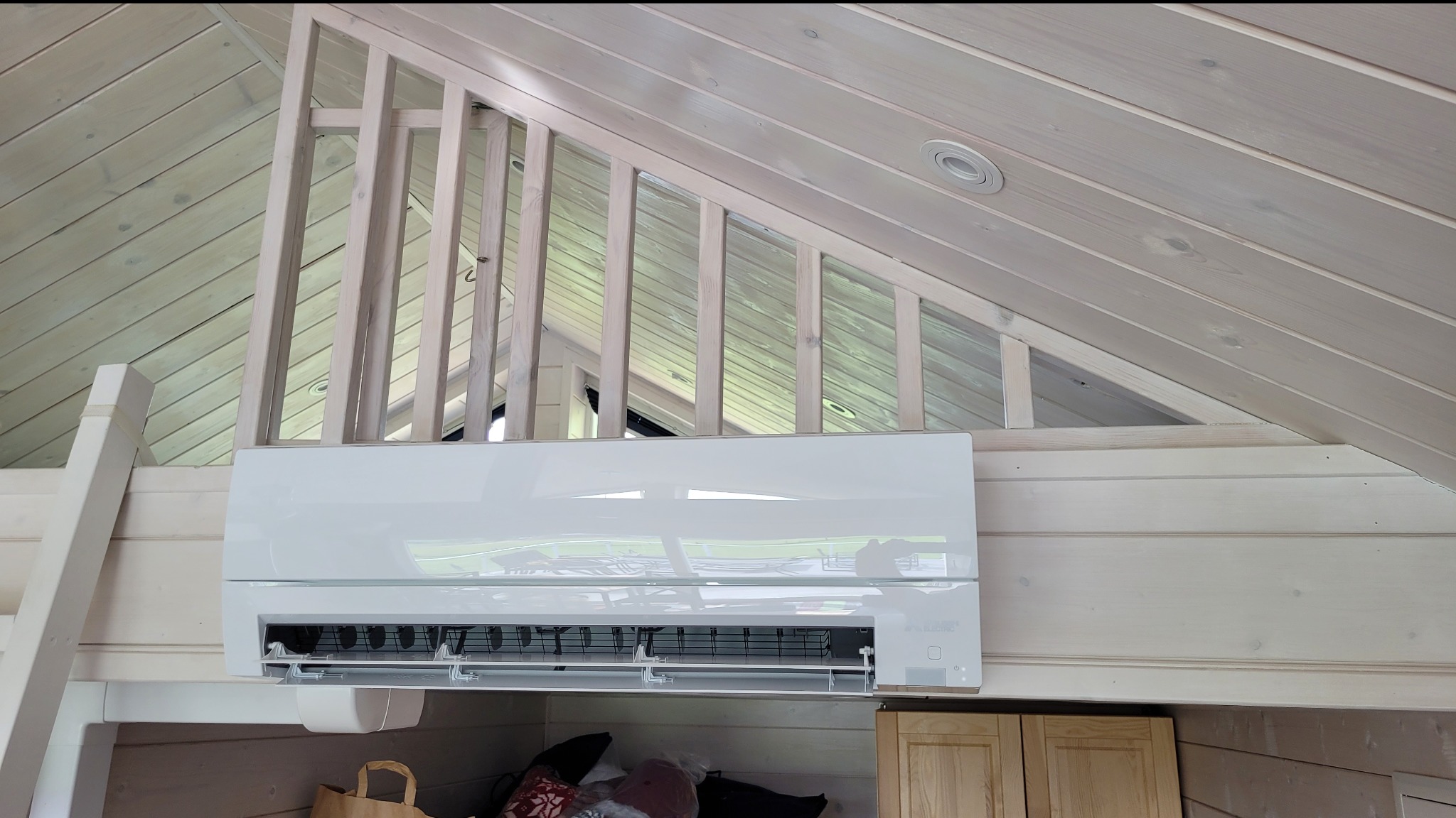 Biały klimatyzator zamontowany w drewnianym domu z antresolą, widoczne drewniane ściany, sufit i barierka antresoli.