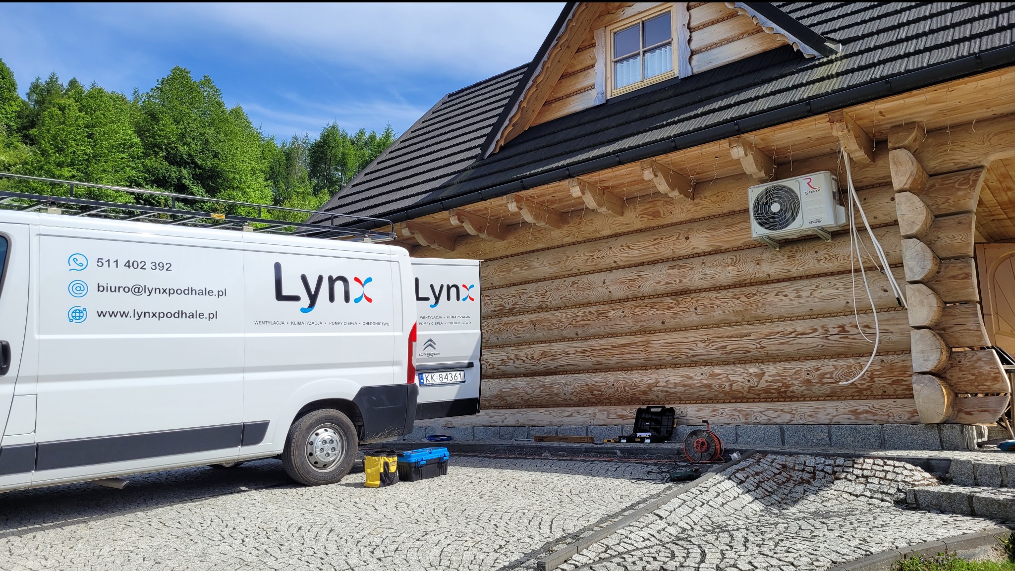 Montaż jednostki zewnętrznej klimatyzacji na drewnianej ścianie domu z bali, widoczny firmowy van z logo Lynx i danymi kontaktowymi, skrzynki narzędziowe obok schodów.