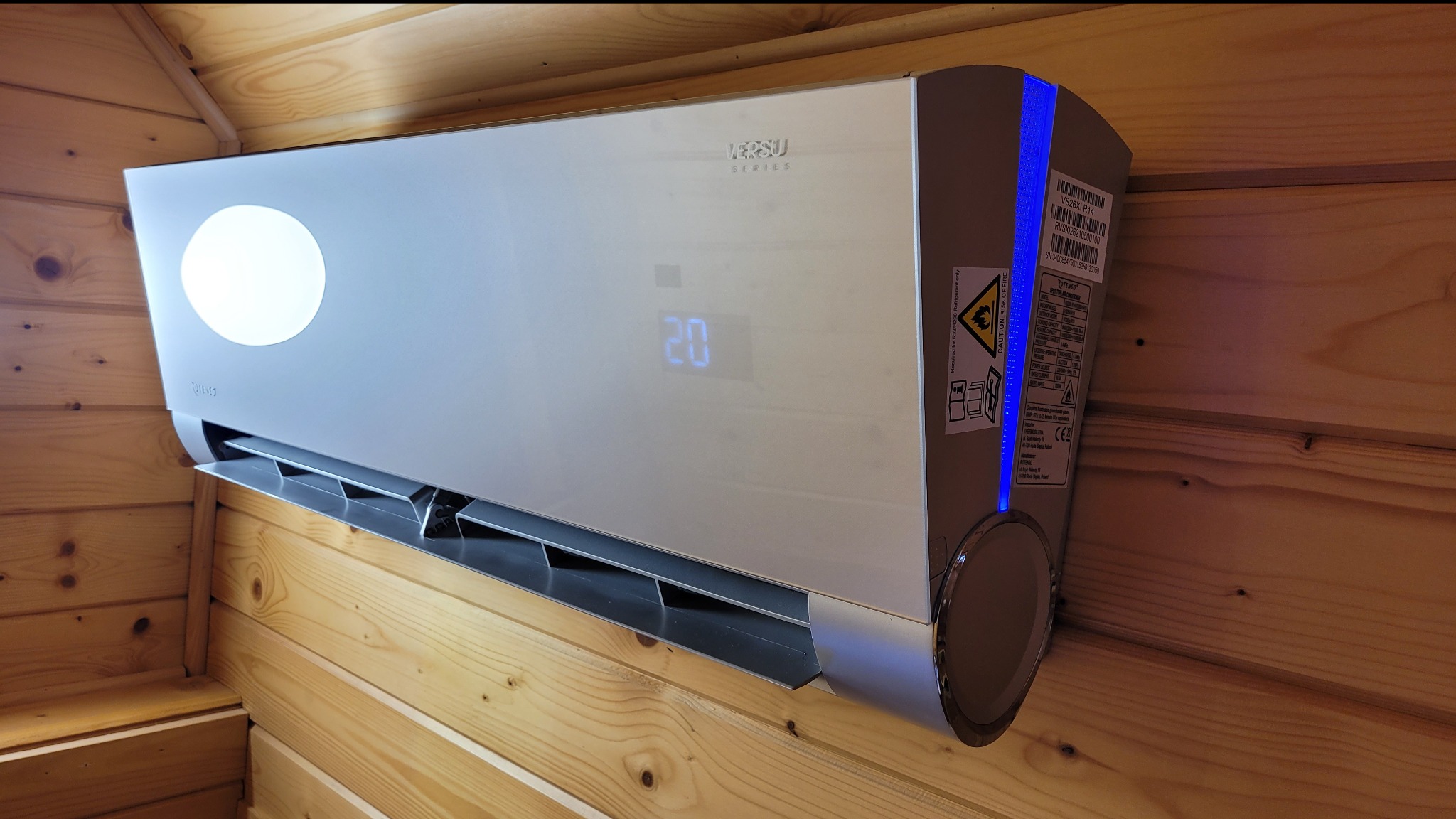 Nowoczesny klimatyzator Versu Series z wyświetlaczem temperatury 20 stopni, zamontowany na drewnianej ścianie w Kluszkowcach, z widocznymi naklejkami informacyjnymi na boku urządzenia.