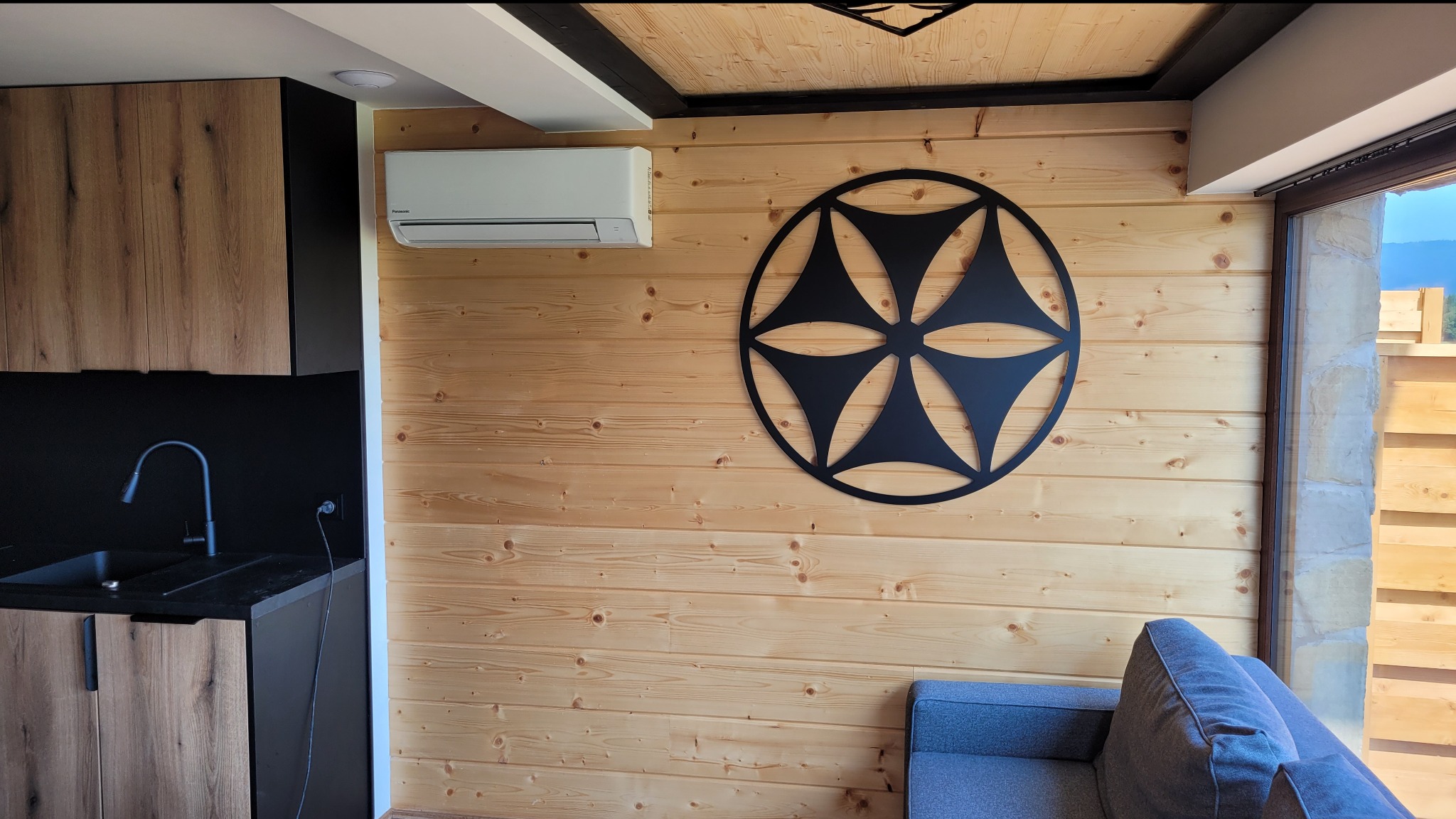 Wnętrze domu z drewnianą boazerią, klimatyzatorem ściennym, dekoracyjną metalową rozetą i fragmentem szarej sofy.