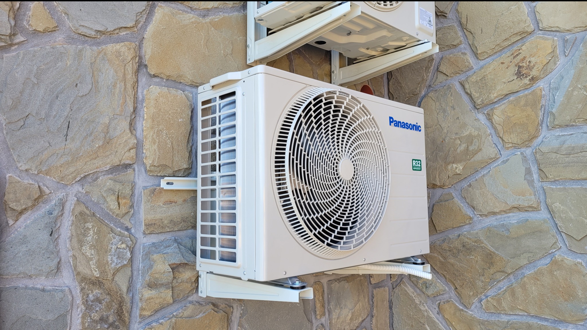 Zewnętrzna jednostka klimatyzatora Panasonic zamontowana na ścianie z kamienia naturalnego, widoczny przewód odprowadzający skropliny i górna jednostka w tle.