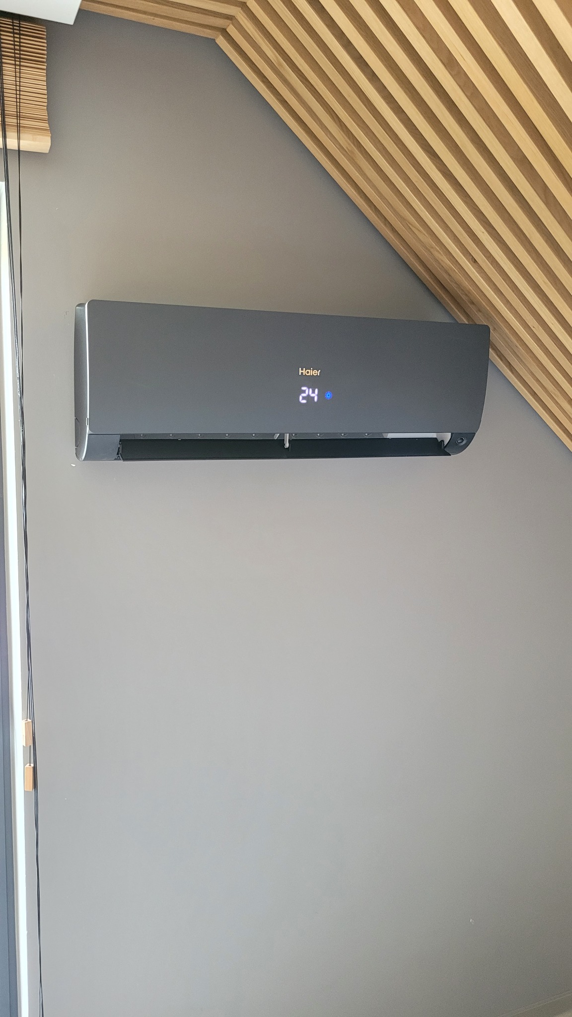 Szary klimatyzator Haier zamontowany na szarej ścianie w pomieszczeniu ze skośnym sufitem wykończonym drewnianymi listwami. Na wyświetlaczu widoczna temperatura 24 stopnie.