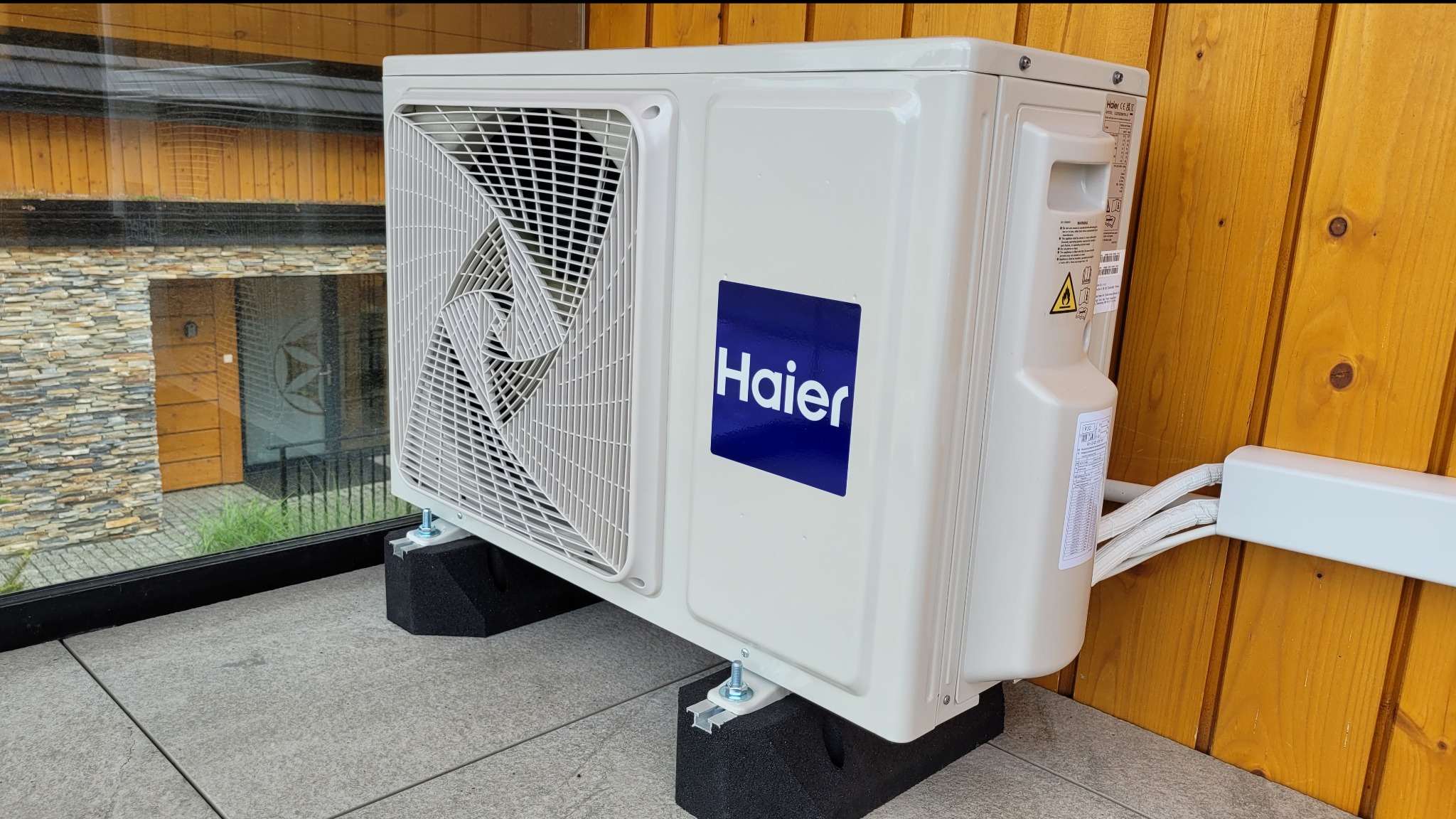 Zewnętrzna jednostka klimatyzatora Haier zamontowana na czarnych podkładkach tłumiących drgania, widoczne przewody w białej listwie, w tle drewniana ściana i okno z widokiem na budynek.